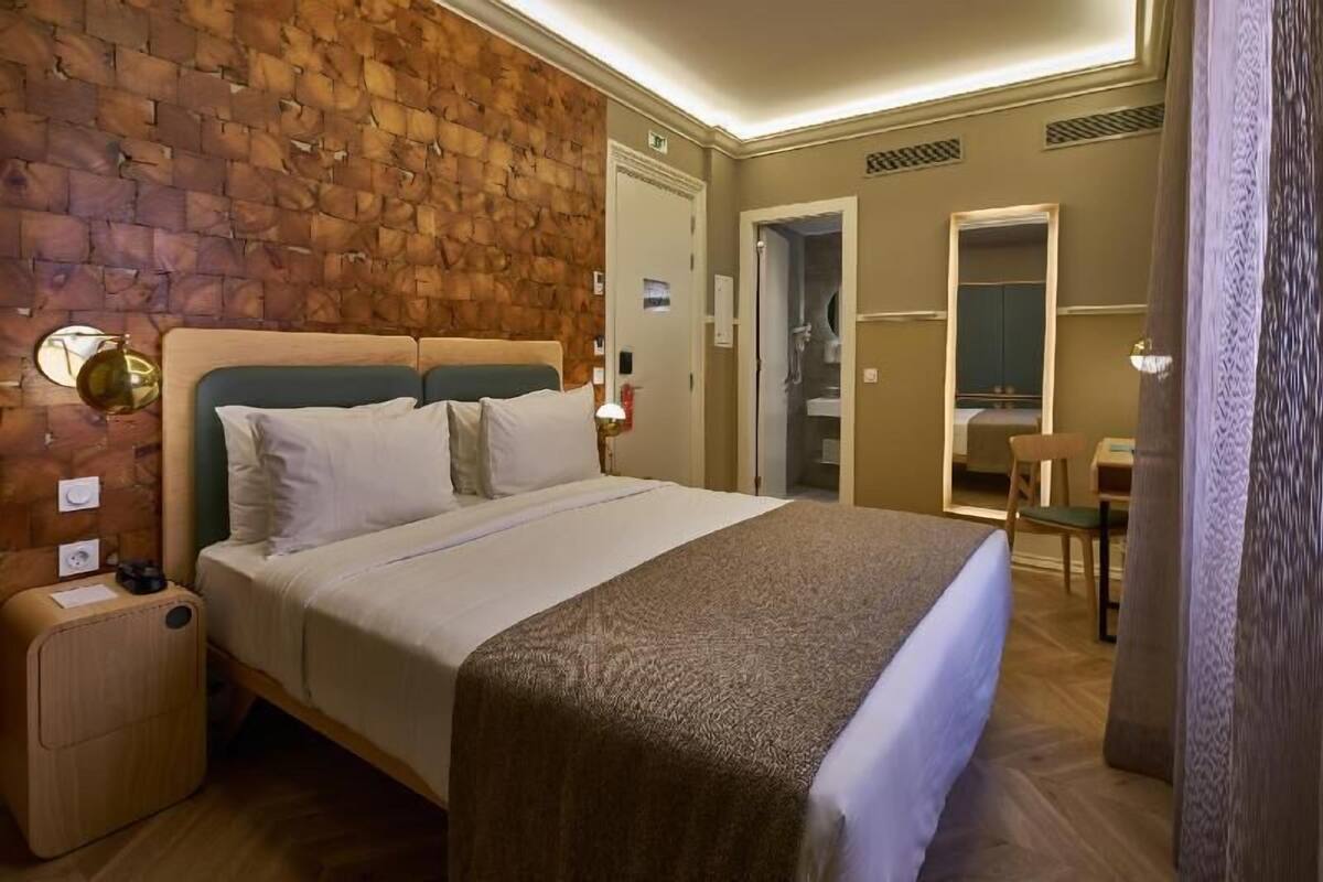 A hotel room with a brick-patterned headboard, a bed with white linens, a small bedside table with a lamp, and a mirror on the wall.