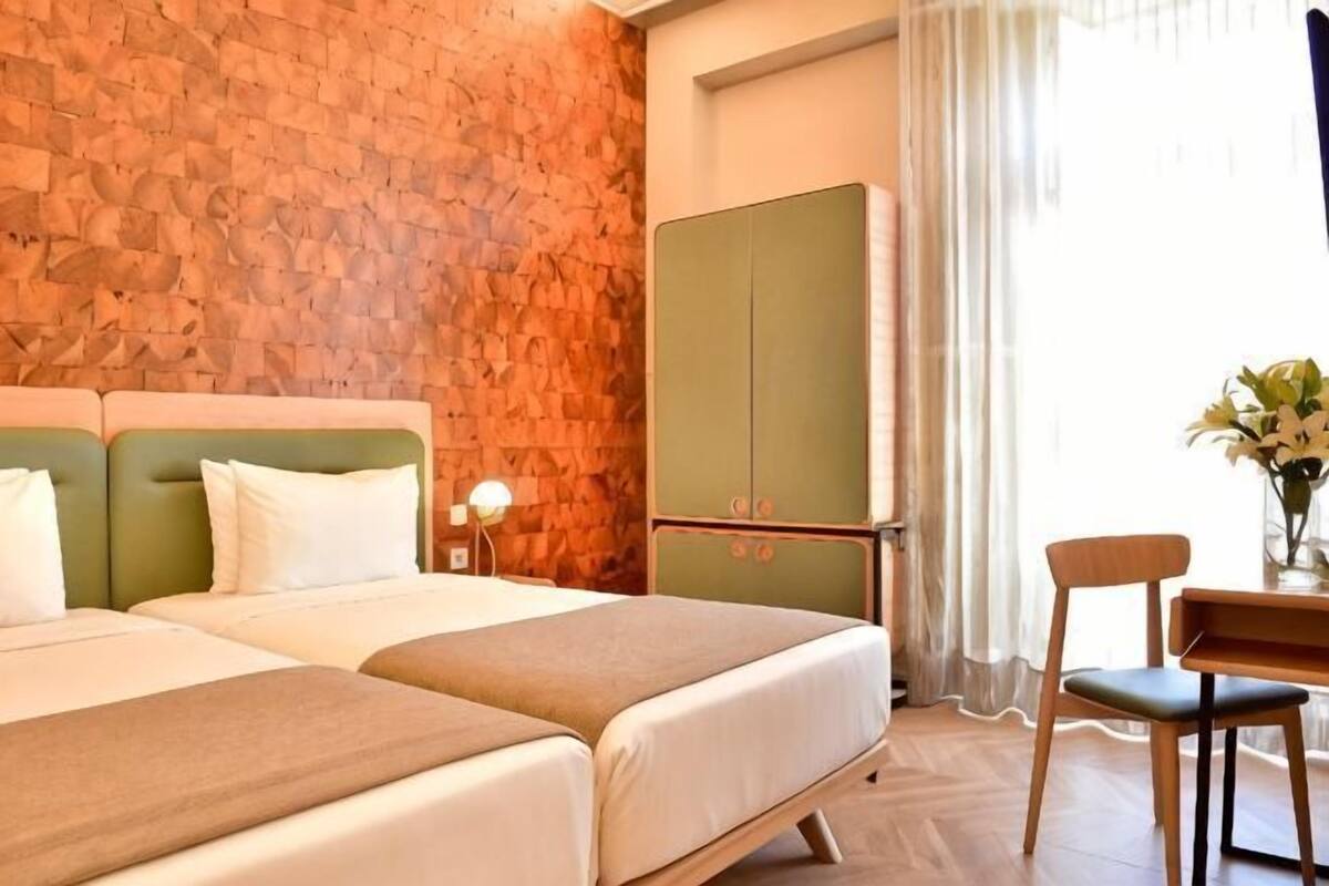 A hotel room with two beds, a green wardrobe, a wooden chair, and a desk with a vase of flowers.