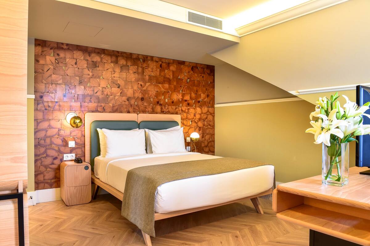 A modern hotel room with a wooden headboard, a large bed, a bedside table with a lamp, and a vase of flowers.