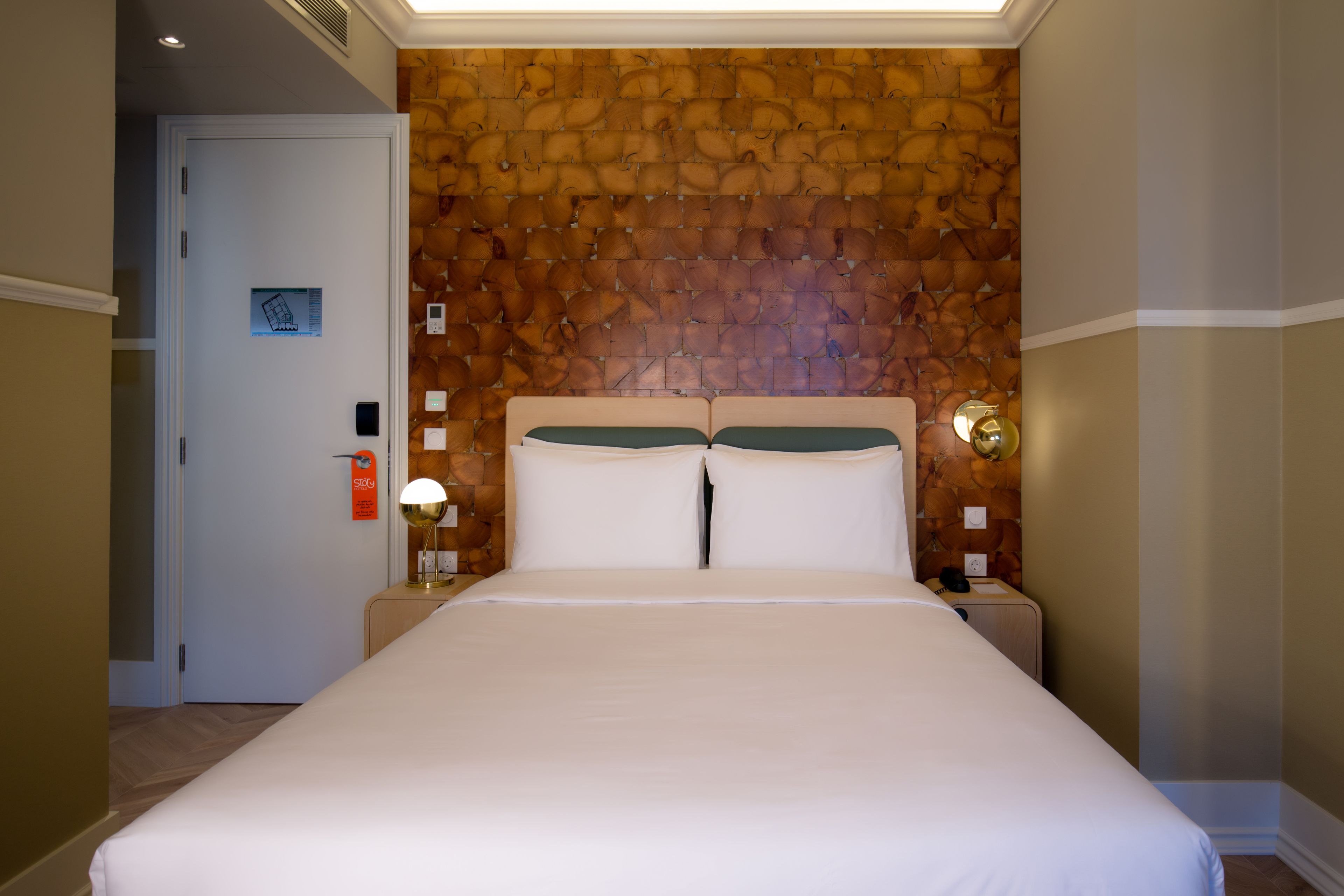A hotel room with a large bed, a wooden headboard, a nightstand with a lamp, and a wall-mounted thermostat.
