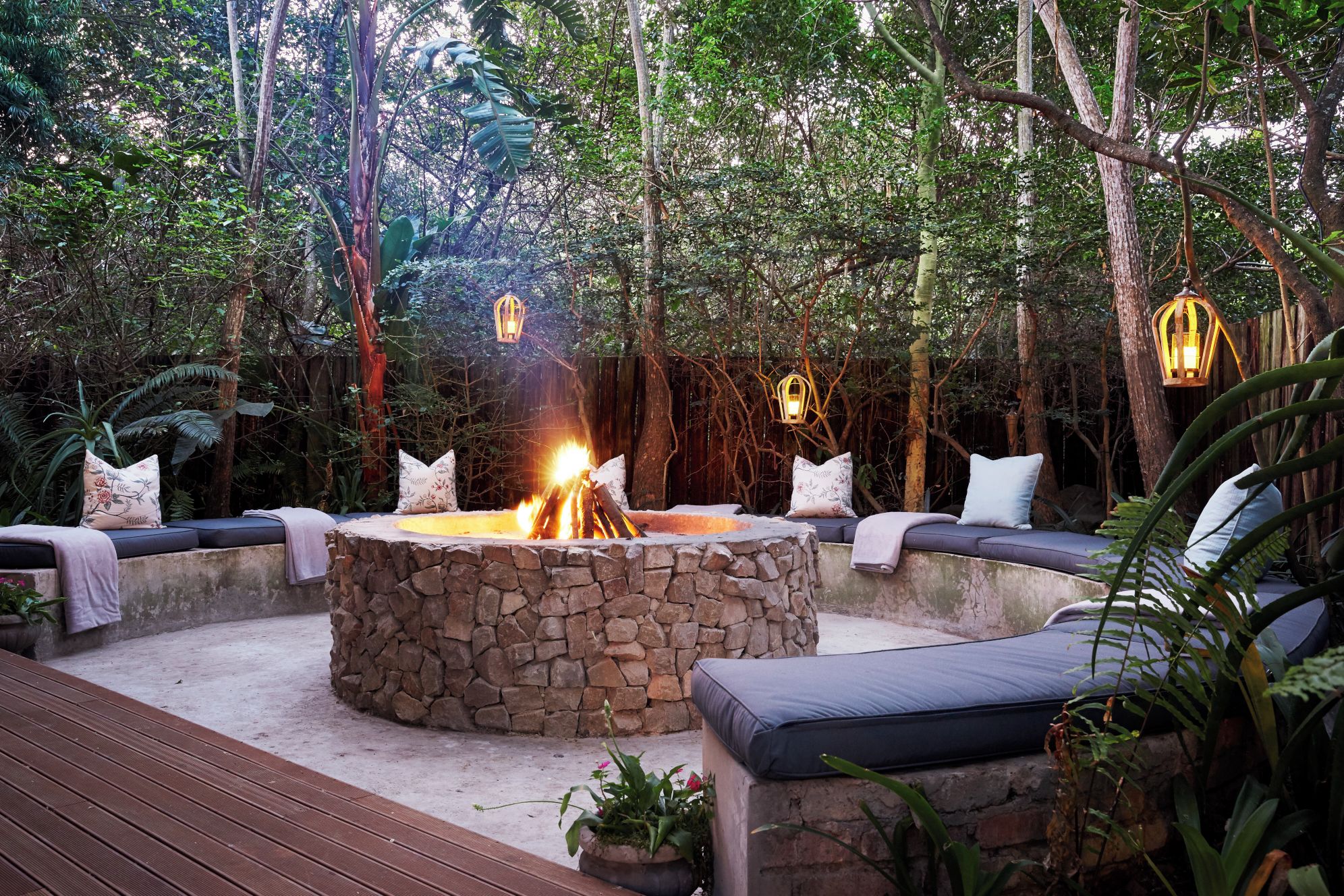 An outdoor seating area with stone fire pit, cushioned benches, and hanging lanterns.