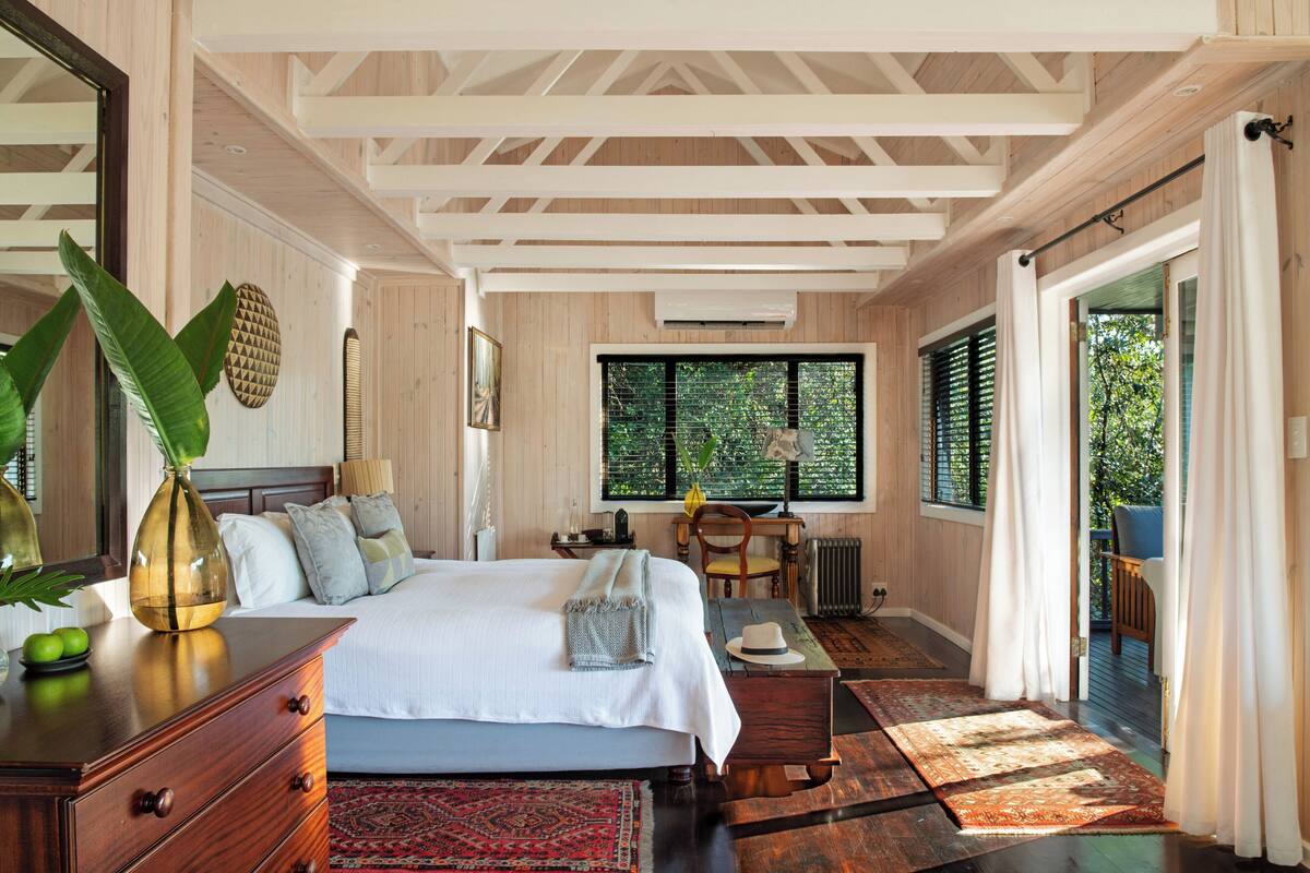 A spacious bedroom with a large bed, wooden ceiling beams, a view through the window, and a wooden floor.