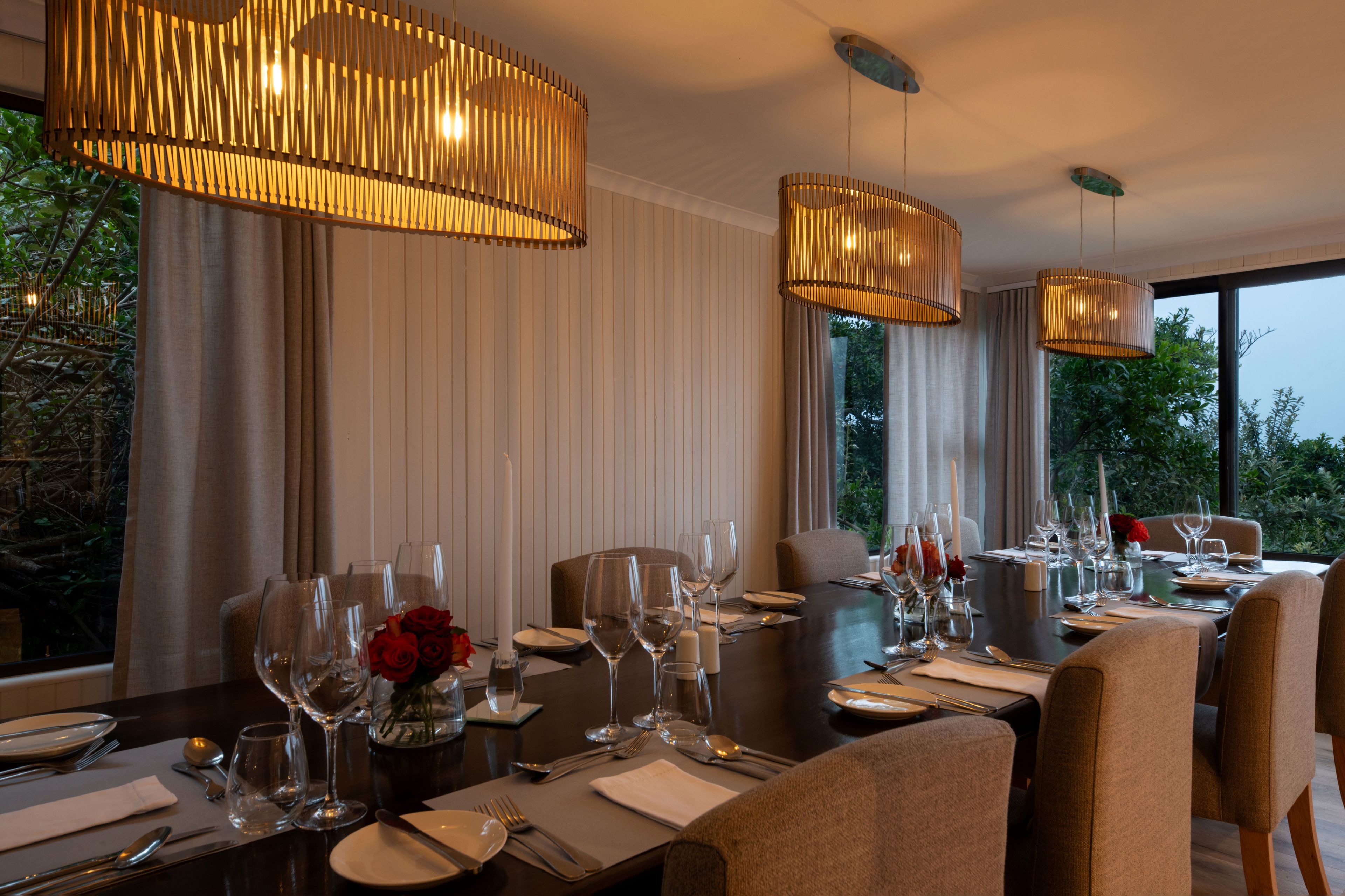 A dining area with a long table set for a meal, featuring multiple chairs, wine glasses, and a centerpiece of red roses.
