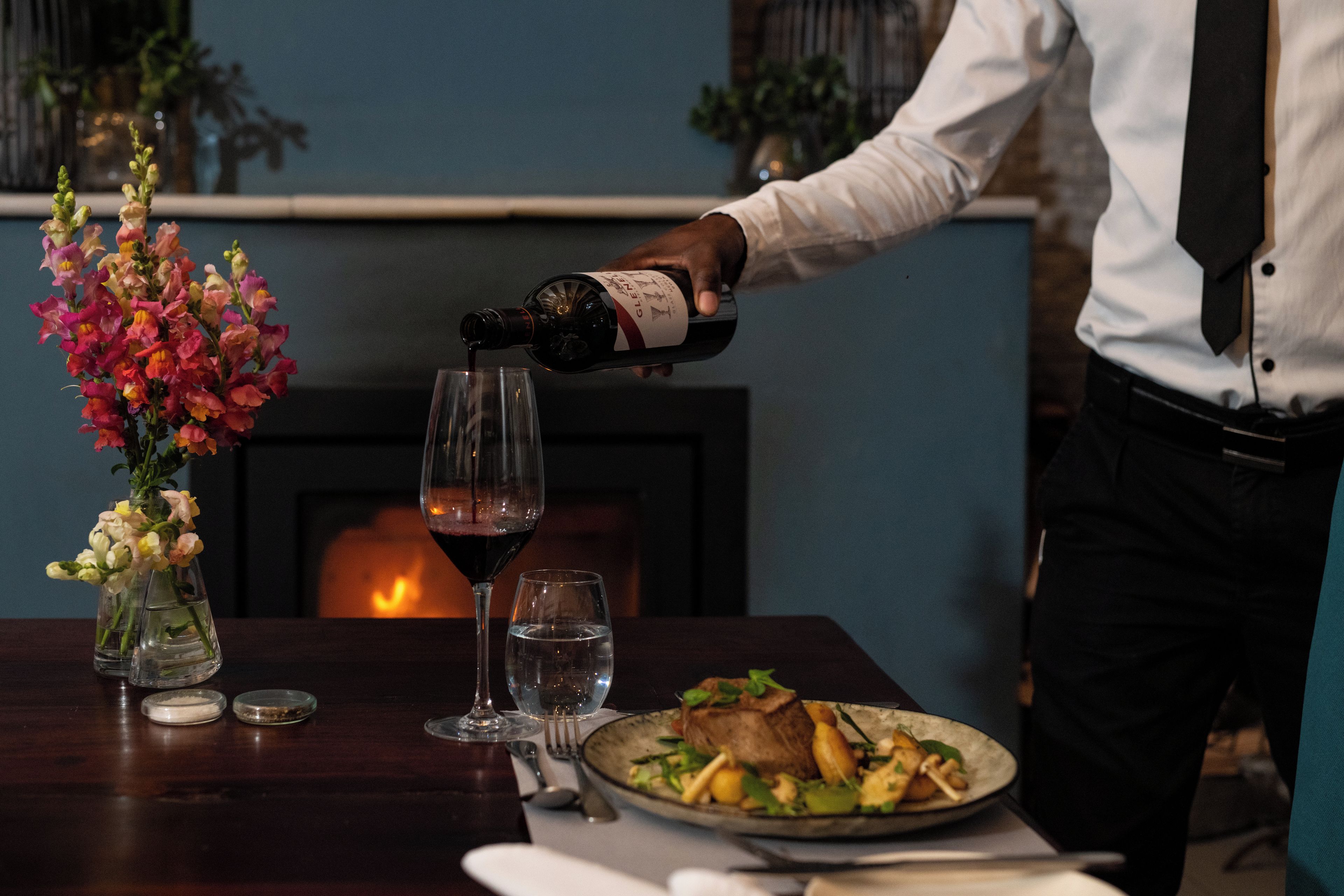 A person pouring a drink into a glass, a vase with flowers, and a plate of food on a table.
