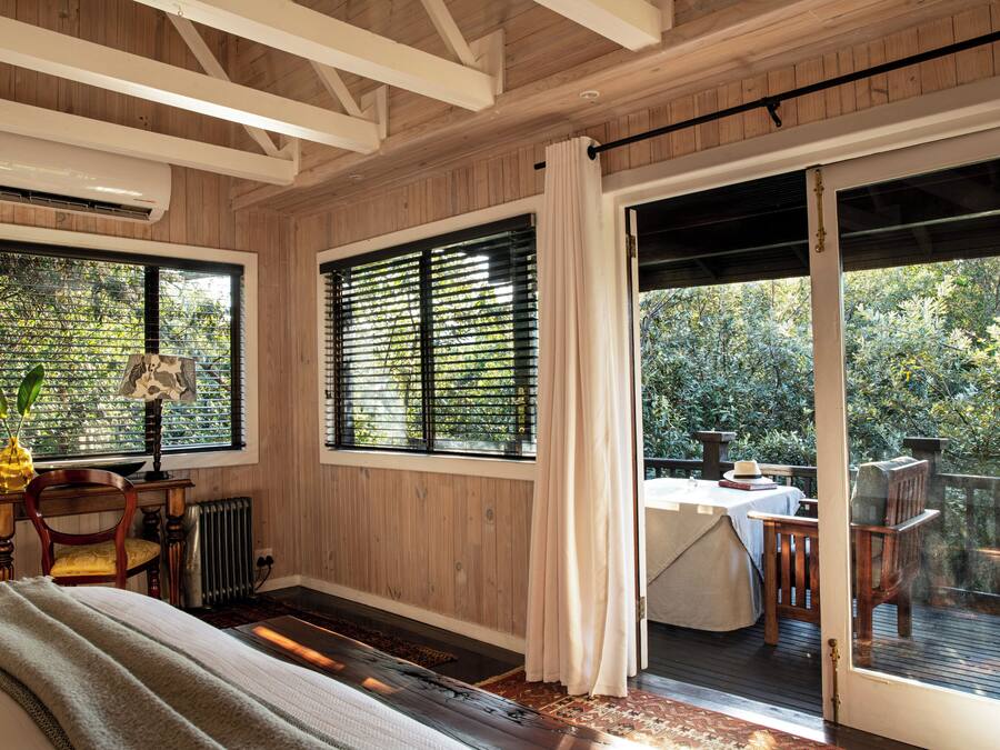 Ein Schlafzimmer mit einem großen Bett, Holzwänden, einem Fenster mit Jalousien, einem Heizkörper und Blick ins Freie.