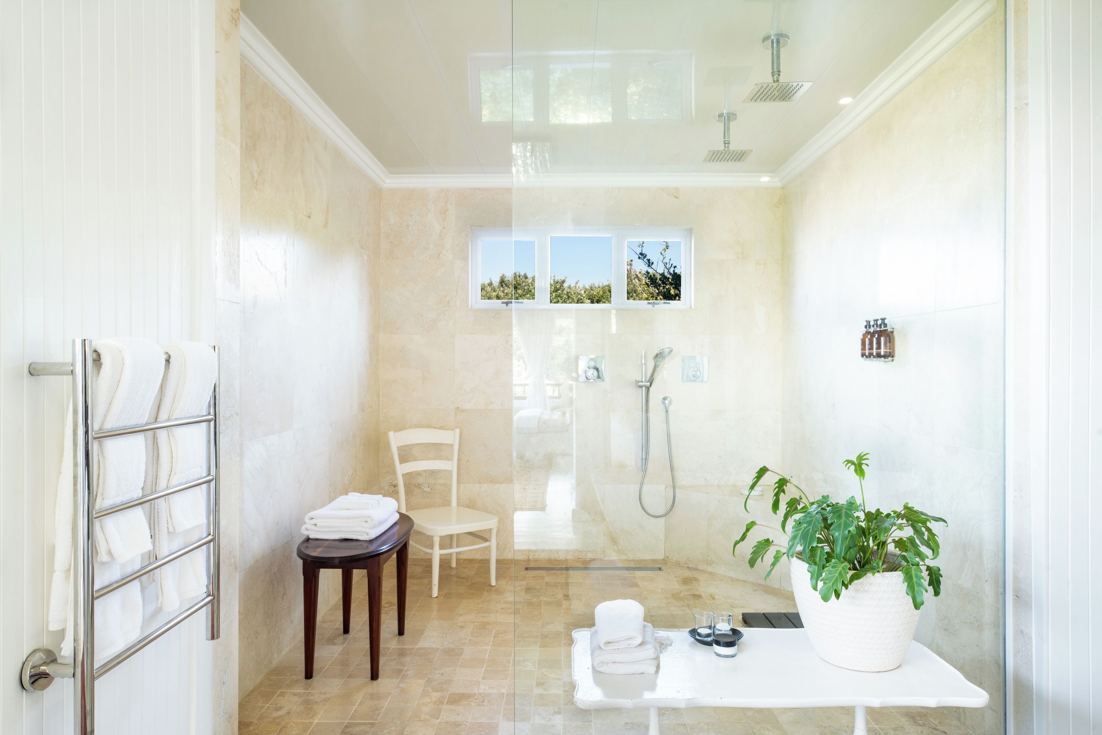 A modern bathroom with a glass shower enclosure, a chair, a table with toiletries, and a potted plant.