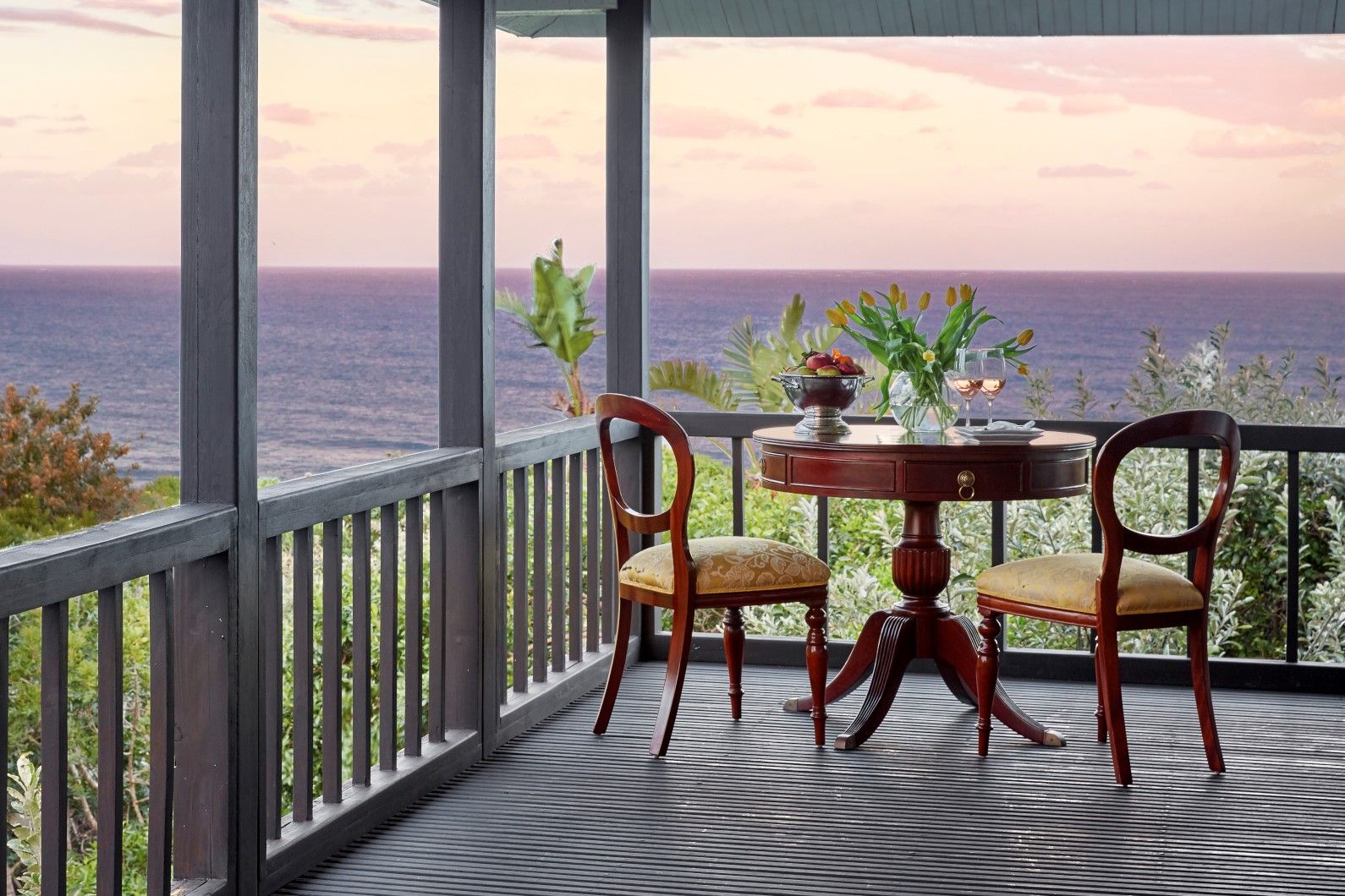 A balcony with a view of the ocean, two wooden chairs, a small round table with a vase and a bowl of fruit.