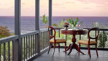 A balcony with a view of the ocean, two wooden chairs, a small round table with a vase and a bowl of fruit.
