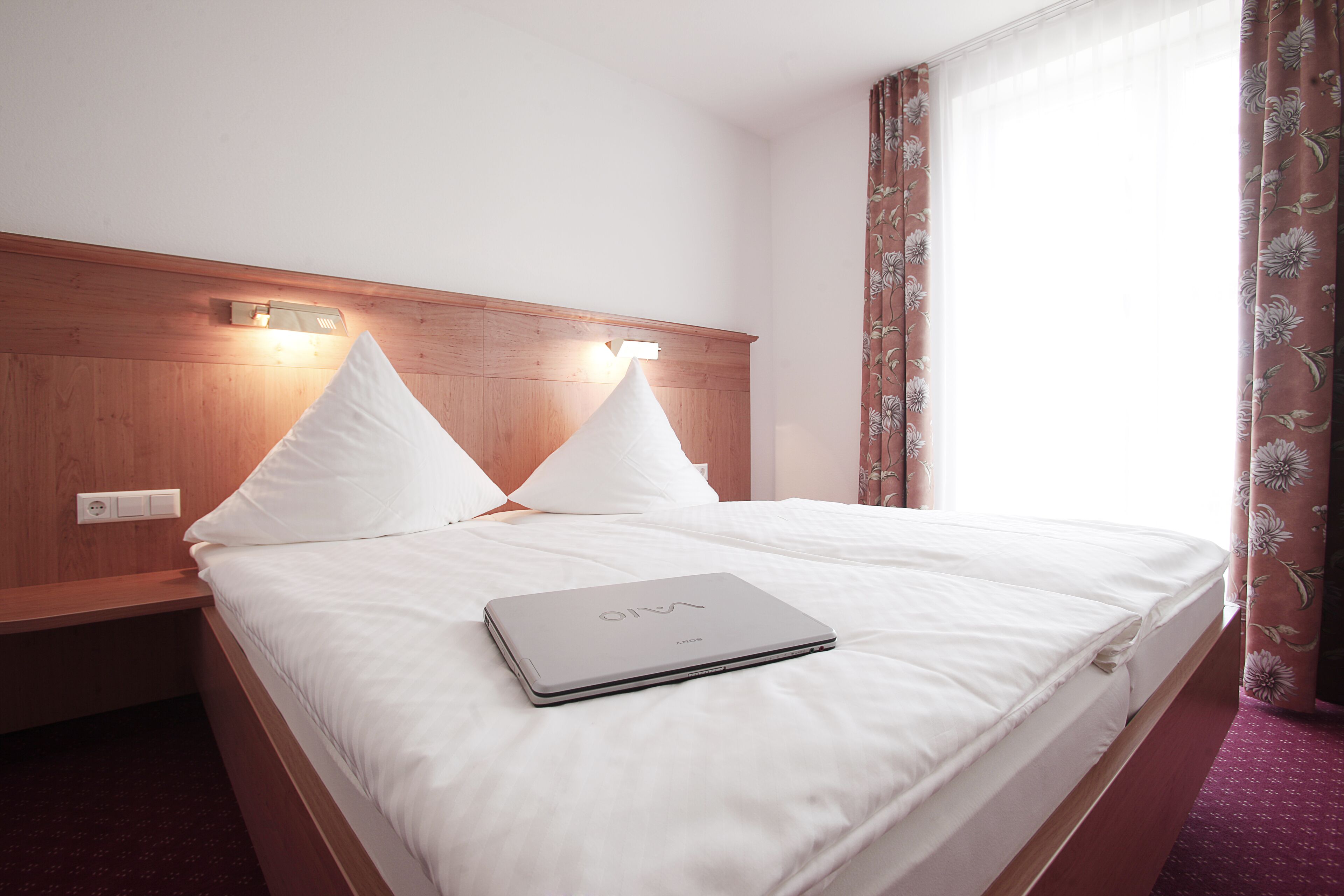 A neatly made bed with white linens and a laptop on top, a wooden headboard, and a window with floral curtains.