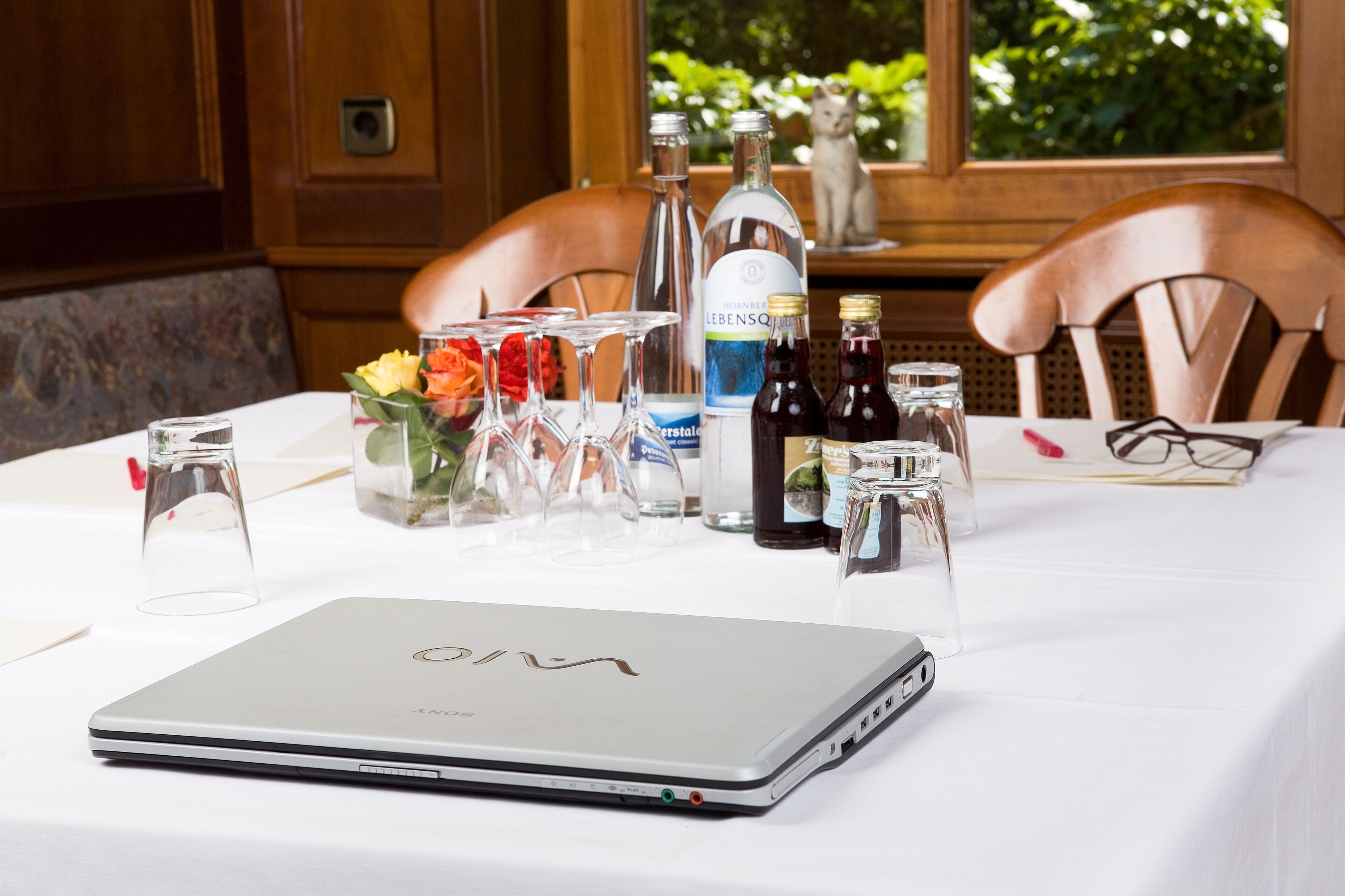 Een tafel met een laptop, wijnflessen, glazen en een vaas met bloemen.