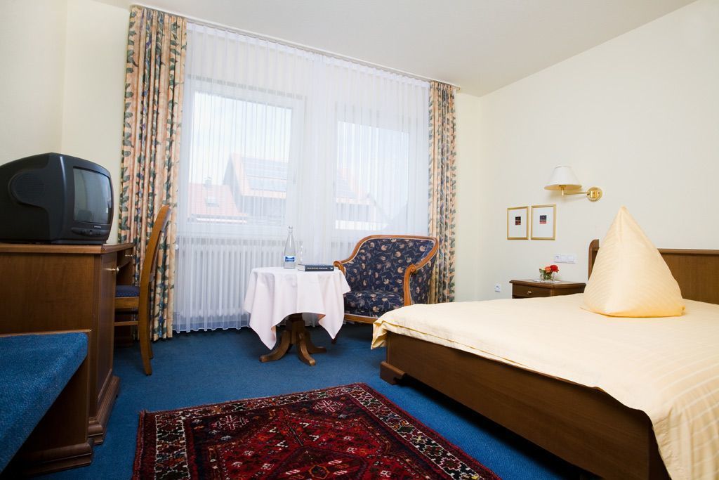 Habitación de hotel con cama, escritorio con televisor, silla, mesita con un libro y ventana con cortinas.