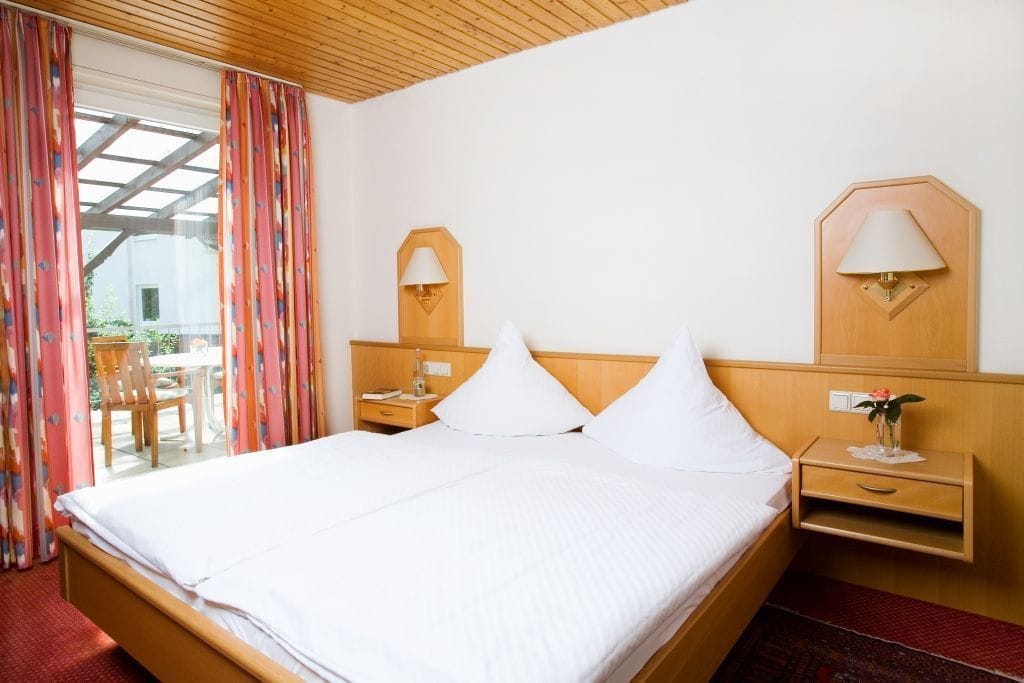 Ein Hotelzimmer mit einem großen Bett, einem hölzernen Kopfteil, zwei Nachttischen mit Lampen, einem kleinen Tisch mit einem Stuhl und Blick durch eine Glastür.