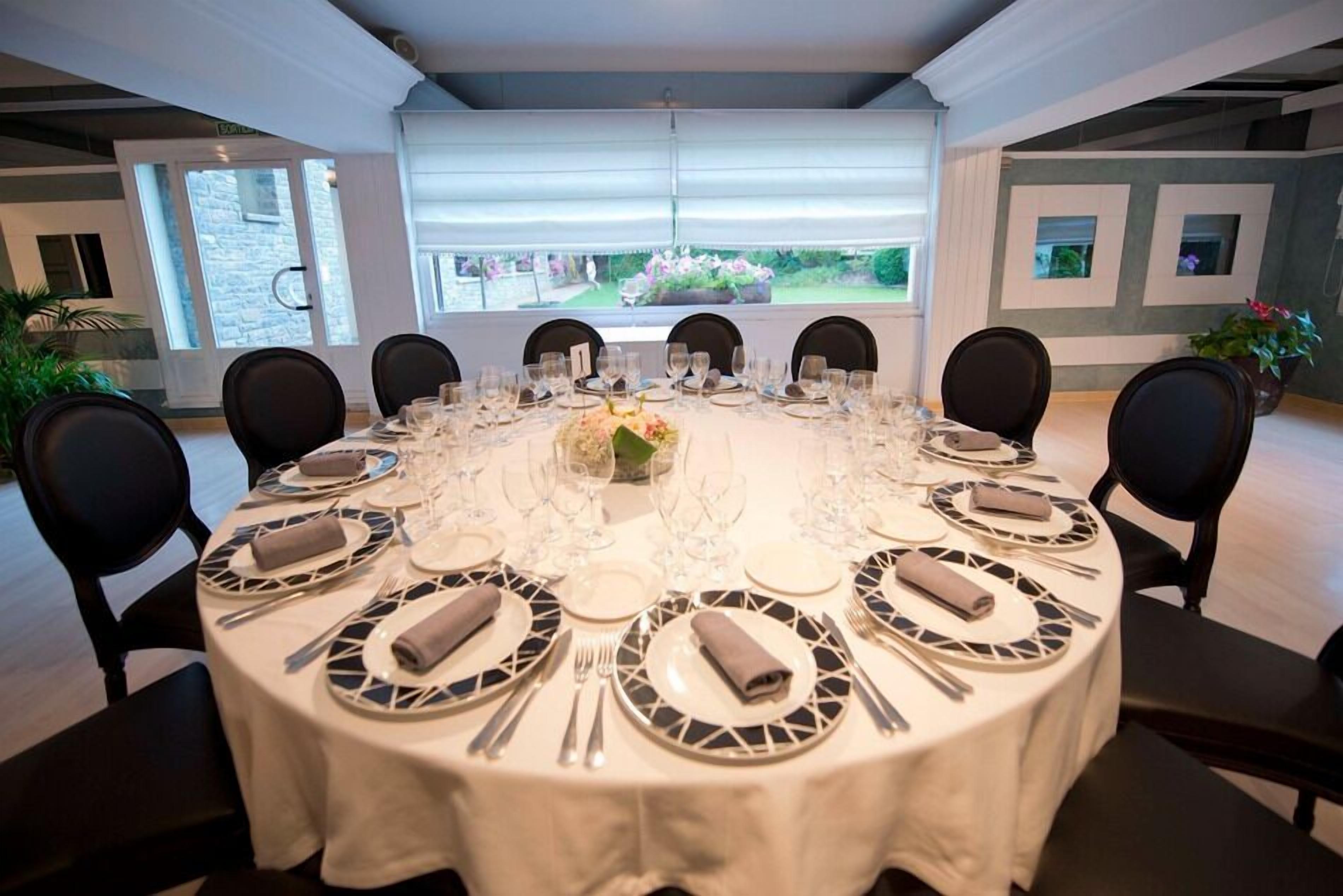 Una mesa de comedor redonda preparada para ocho personas, con mantel blanco, platos blanco y negro y cubiertos.