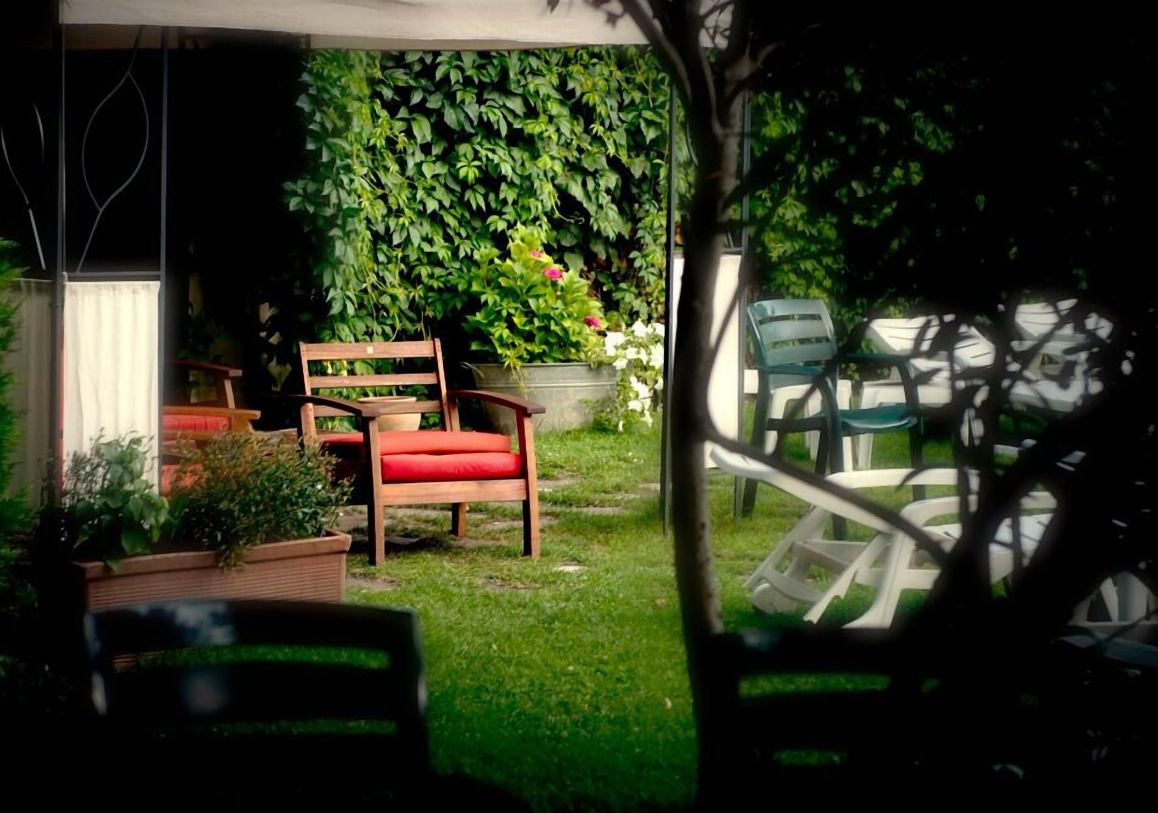 Un patio con un banco de madera y una silla roja, rodeado de abundante vegetación y un dosel blanco.