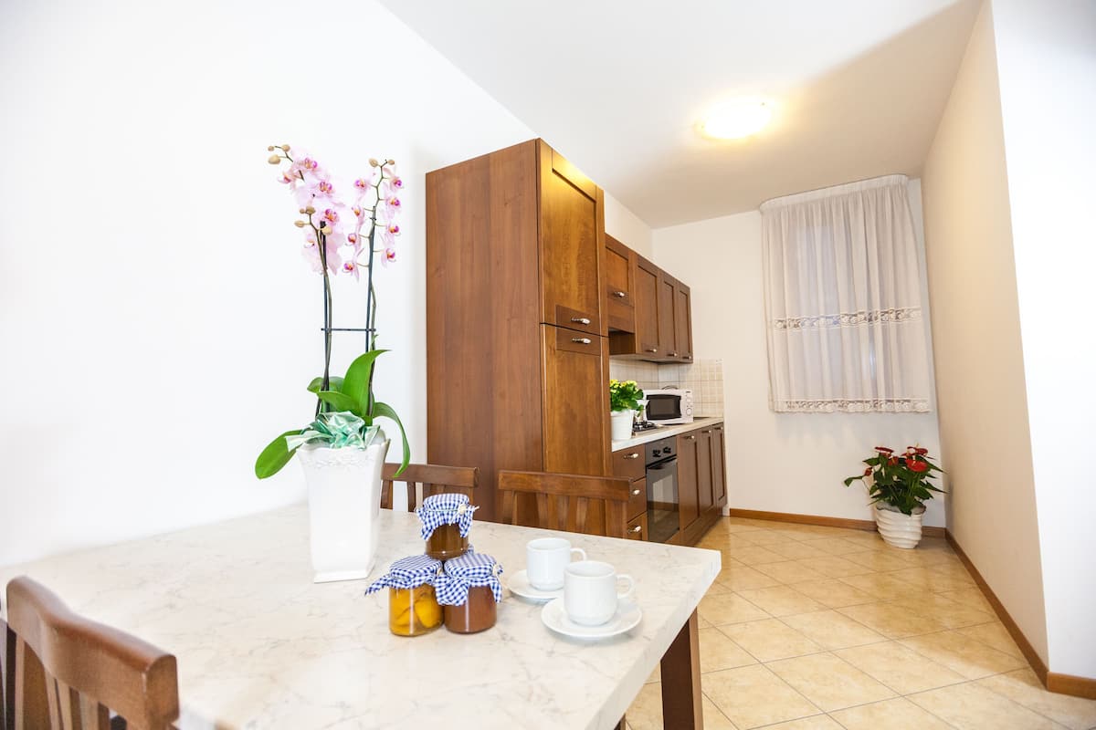 A small kitchen with wooden cabinets, a microwave, and a sink. There is a dining table with a vase of flowers and a jar of jam.
