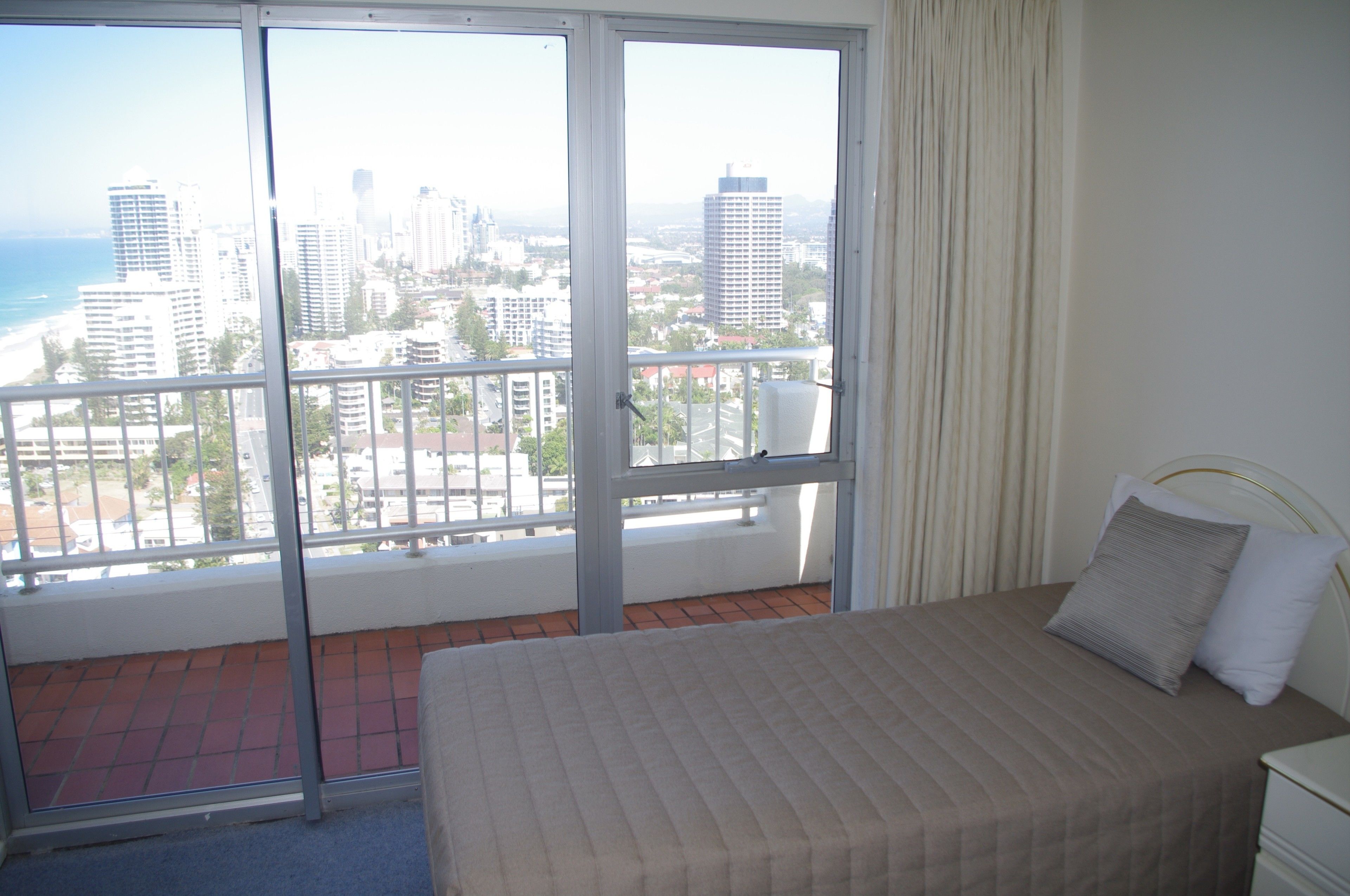 A hotel room with a bed, a nightstand, and a view of a cityscape through large windows.