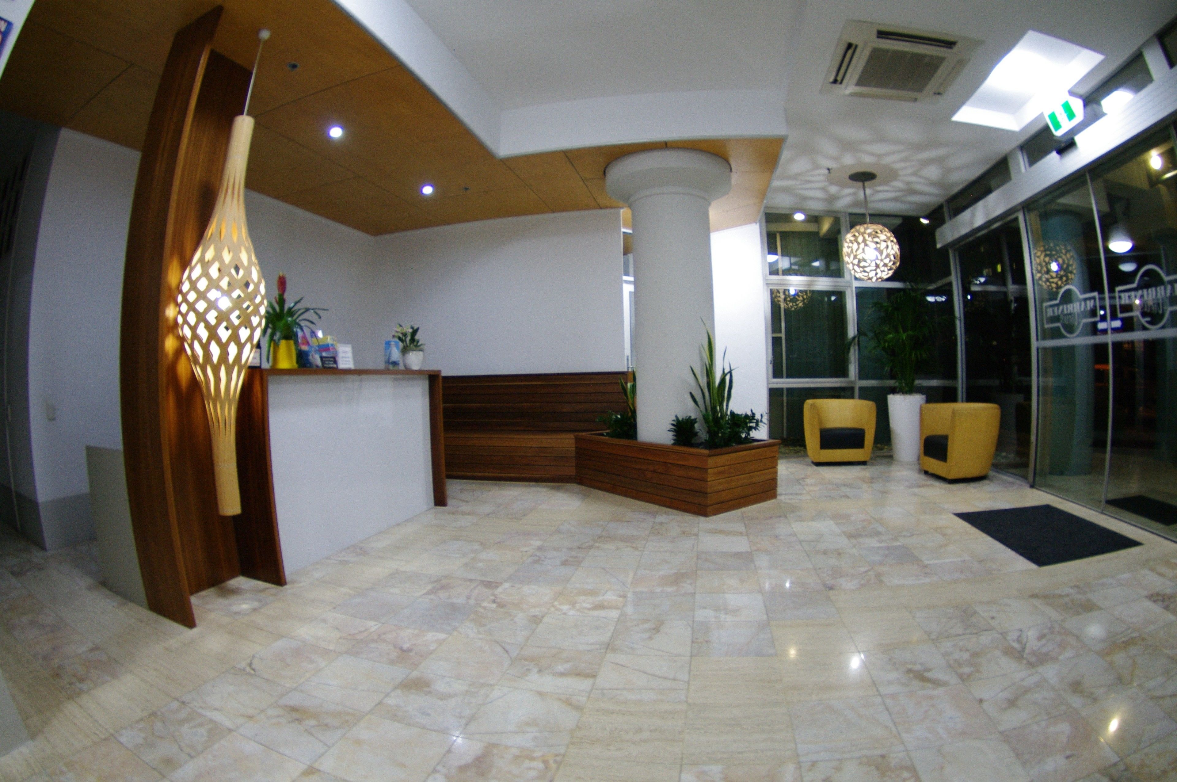 Uma área de recepção com um banco de madeira, um vaso com plantas, uma luminária moderna e uma parede de vidro com reflexos.