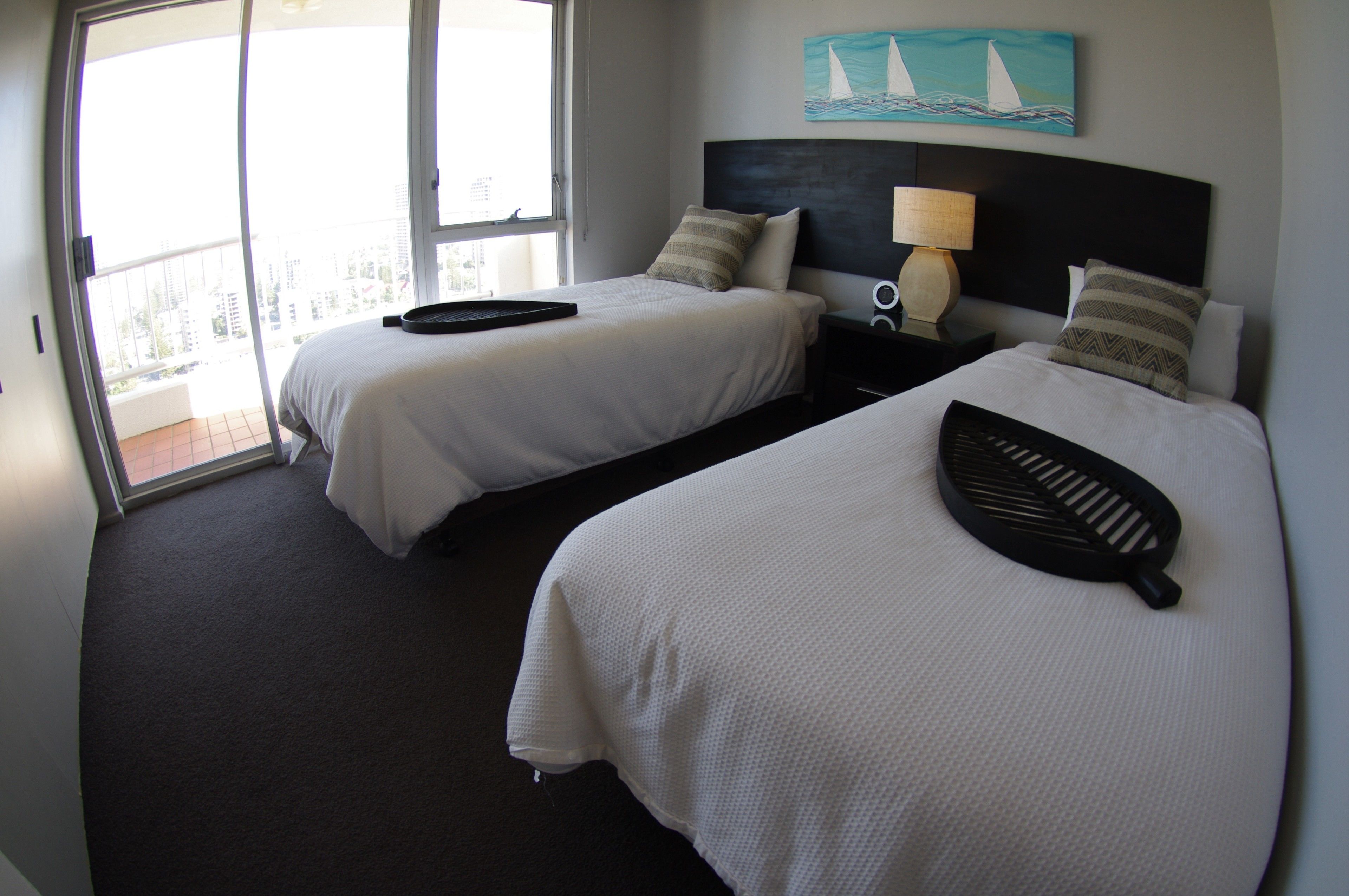 A hotel room with two beds, a painting of sailboats, a lamp, and a view of the city through the balcony doors.