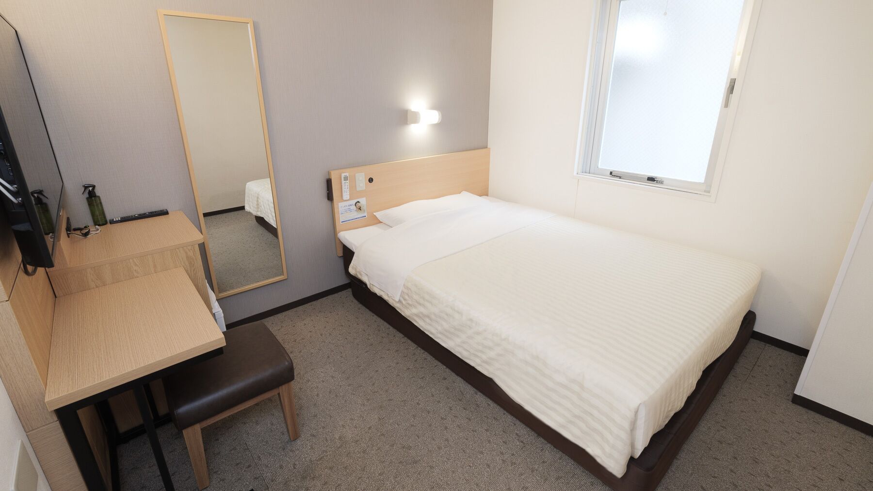 Habitación de hotel con cama, escritorio con silla, espejo, mesita de noche, luz de pared y ventana.