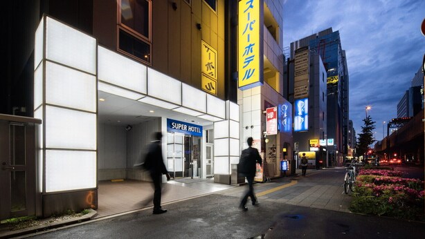 A hotel with a modern facade, a prominent 'Super Hotel' sign, and a pedestrian walkway with flowers.