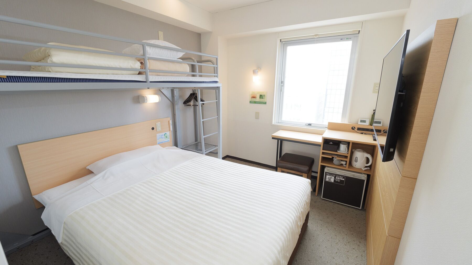 Habitación de hotel con literas, un escritorio con silla, un televisor de pantalla plana y una pequeña kitchenette.