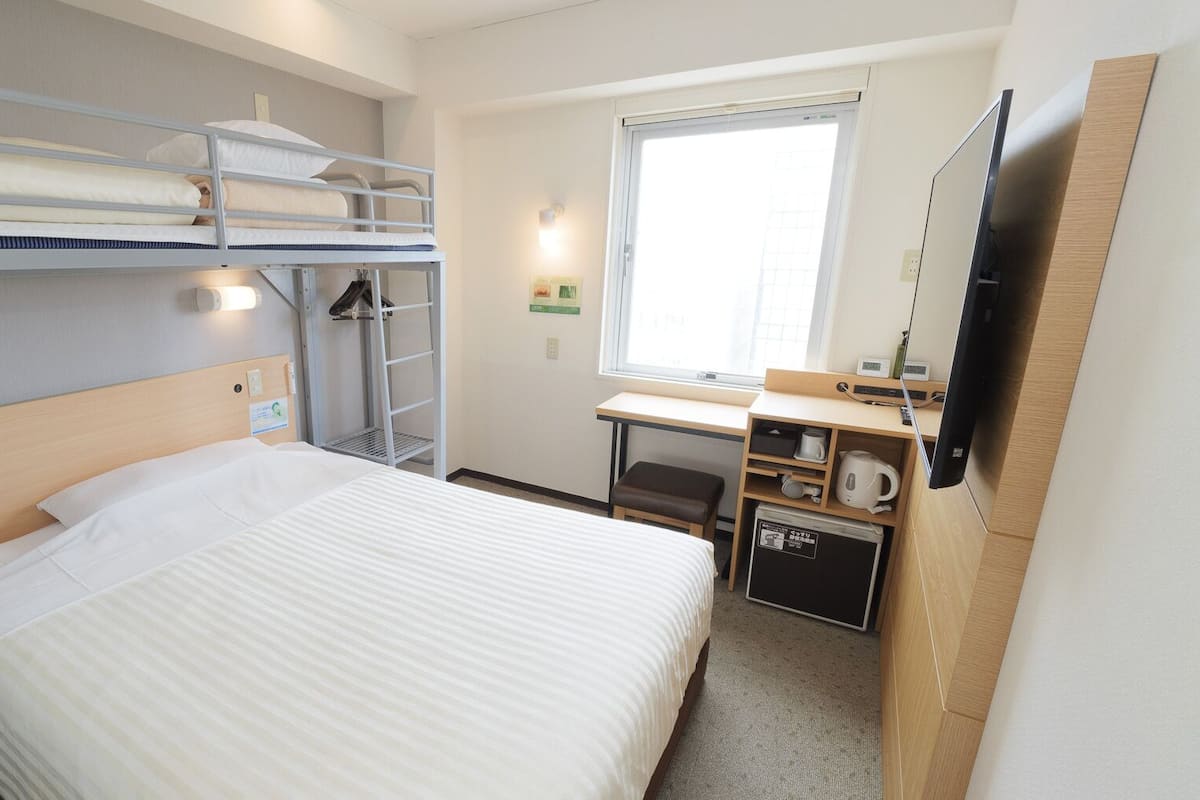 Habitación de hotel con literas, un escritorio con silla, un televisor de pantalla plana y una pequeña kitchenette.