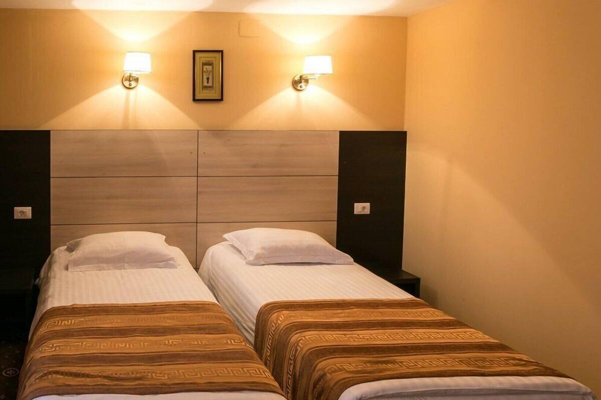 Habitación de hotel con dos camas individuales, cabecero de madera, lámparas fijadas a la pared y un cuadro enmarco.