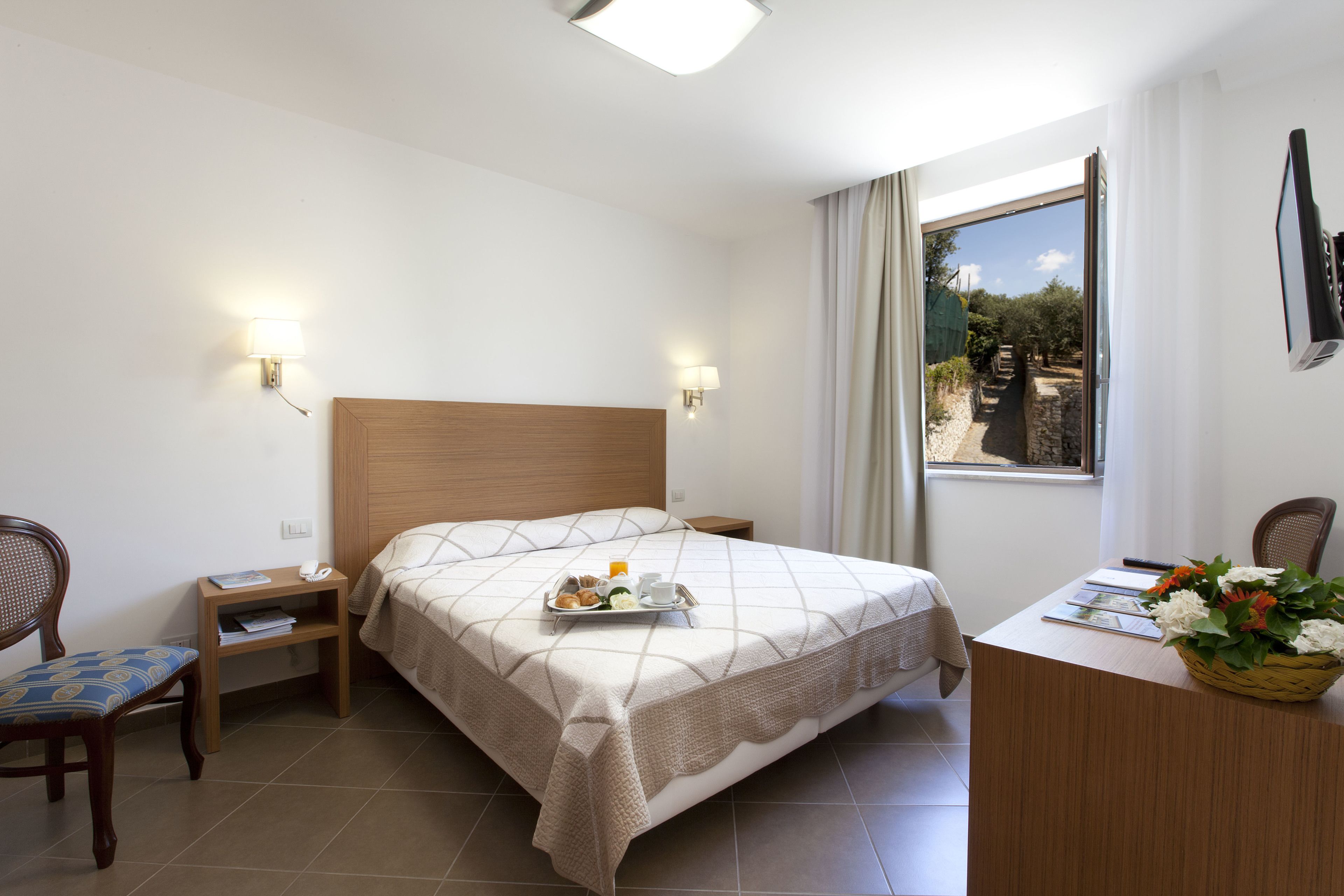 A hotel room with a bed, bedside tables, a desk, a chair, and a window with a view of greenery.