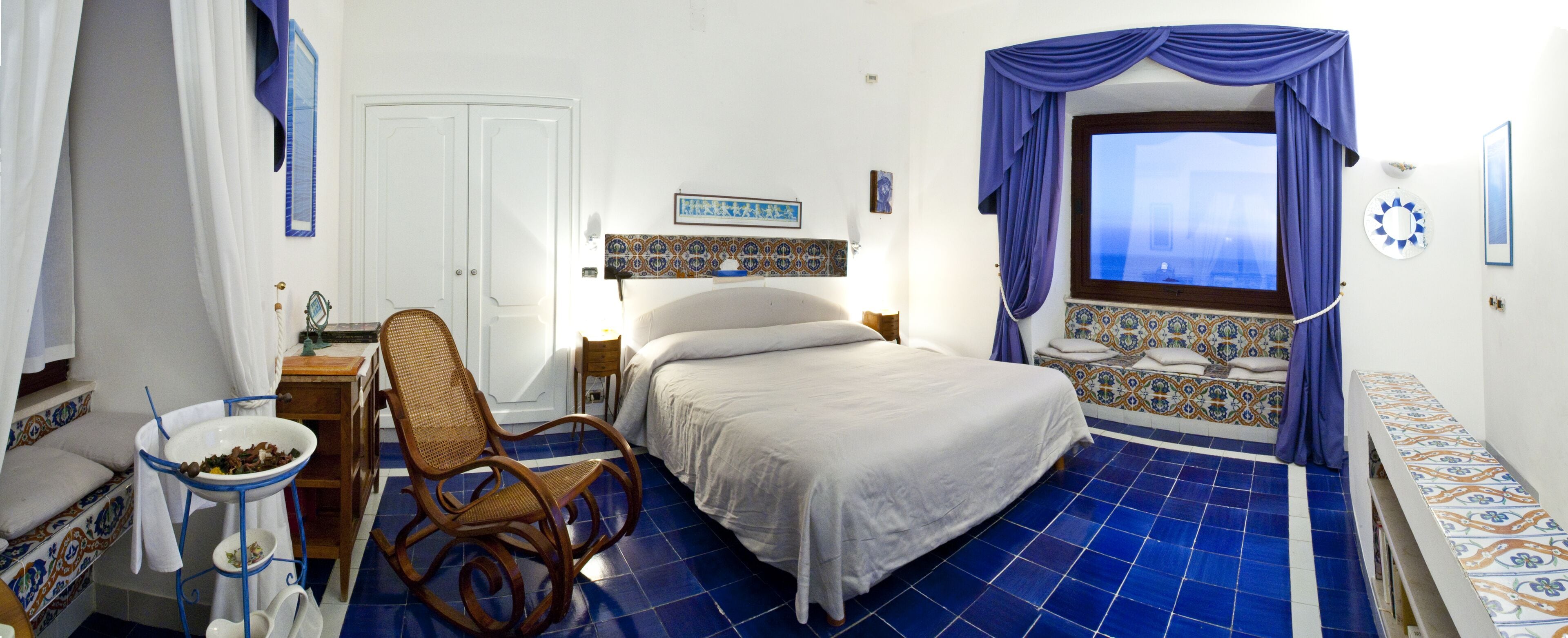 Une chambre à coucher avec un lit, une chaise, une petite table, une fenêtre avec des rideaux et un plancher carrelé.