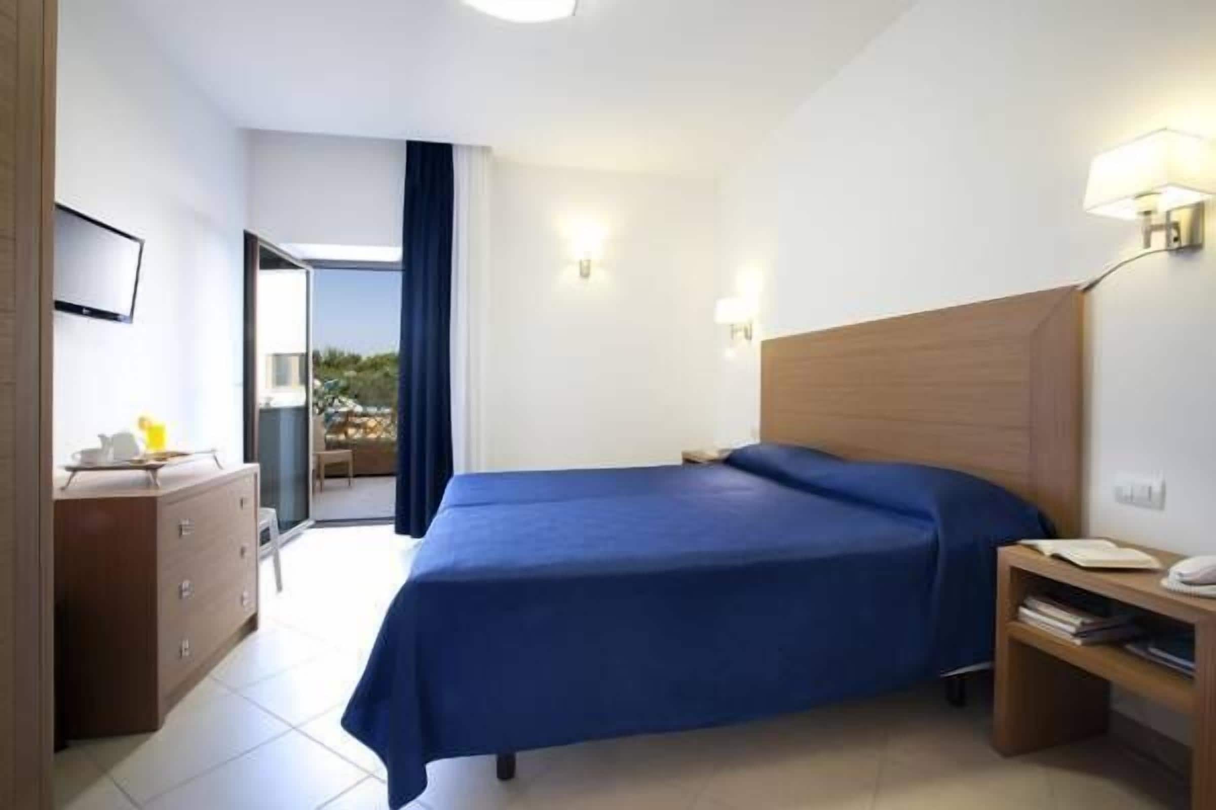 A hotel room with a bed, a wooden nightstand, a desk with a lamp, and a TV on the wall.