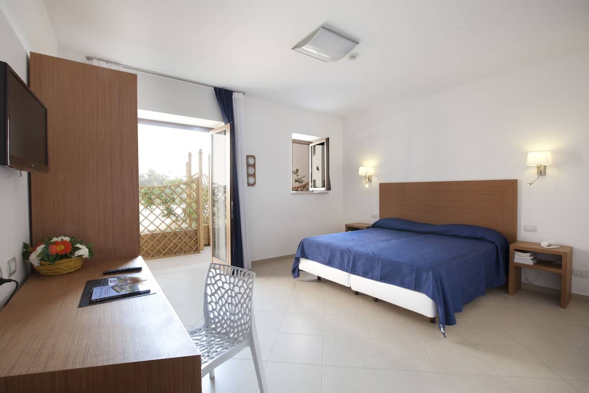 Ein Schlafzimmer mit Bett, Schreibtisch, Stuhl, Nachttisch, Lampe und einem Fenster mit Blick auf Grünflächen.