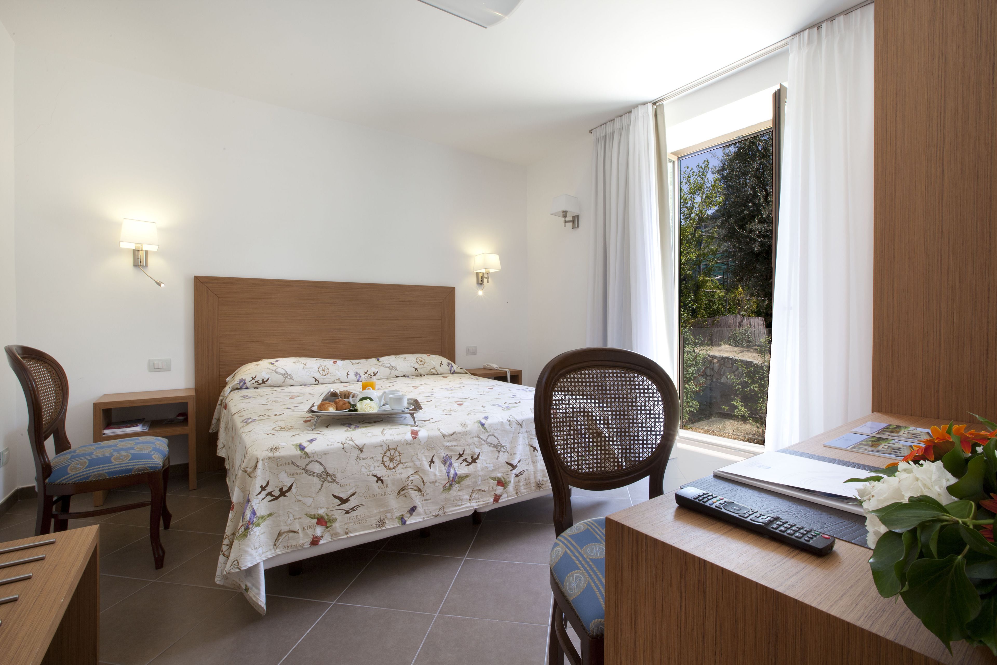 A hotel room with a bed, two chairs, a desk with a laptop and remote, and a window with curtains.