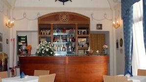 A bar area with a wooden counter, shelves with bottles, and a decorative emblem. There are tables and chairs arranged in front of the bar. - Villa Angelina (Massa Lubrense)