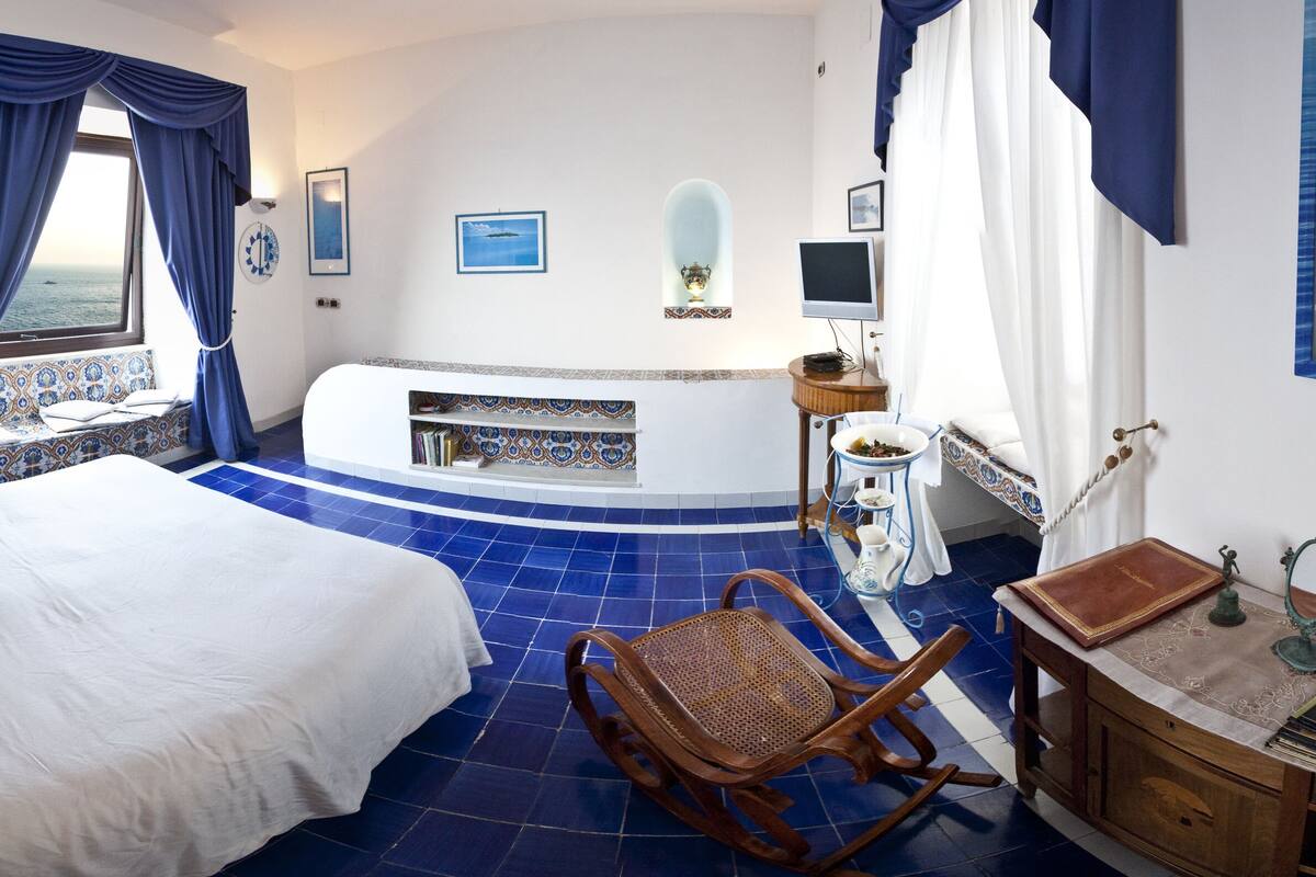 Ein Schlafzimmer mit einem großen Bett, einer Badewanne, einem Sessel, einem Schminktisch mit Spiegel und Meerblick.