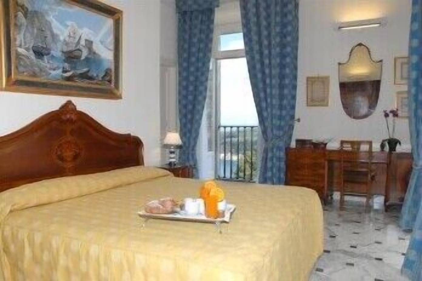 A hotel room with a bed, a painting, a desk, and a window with curtains.