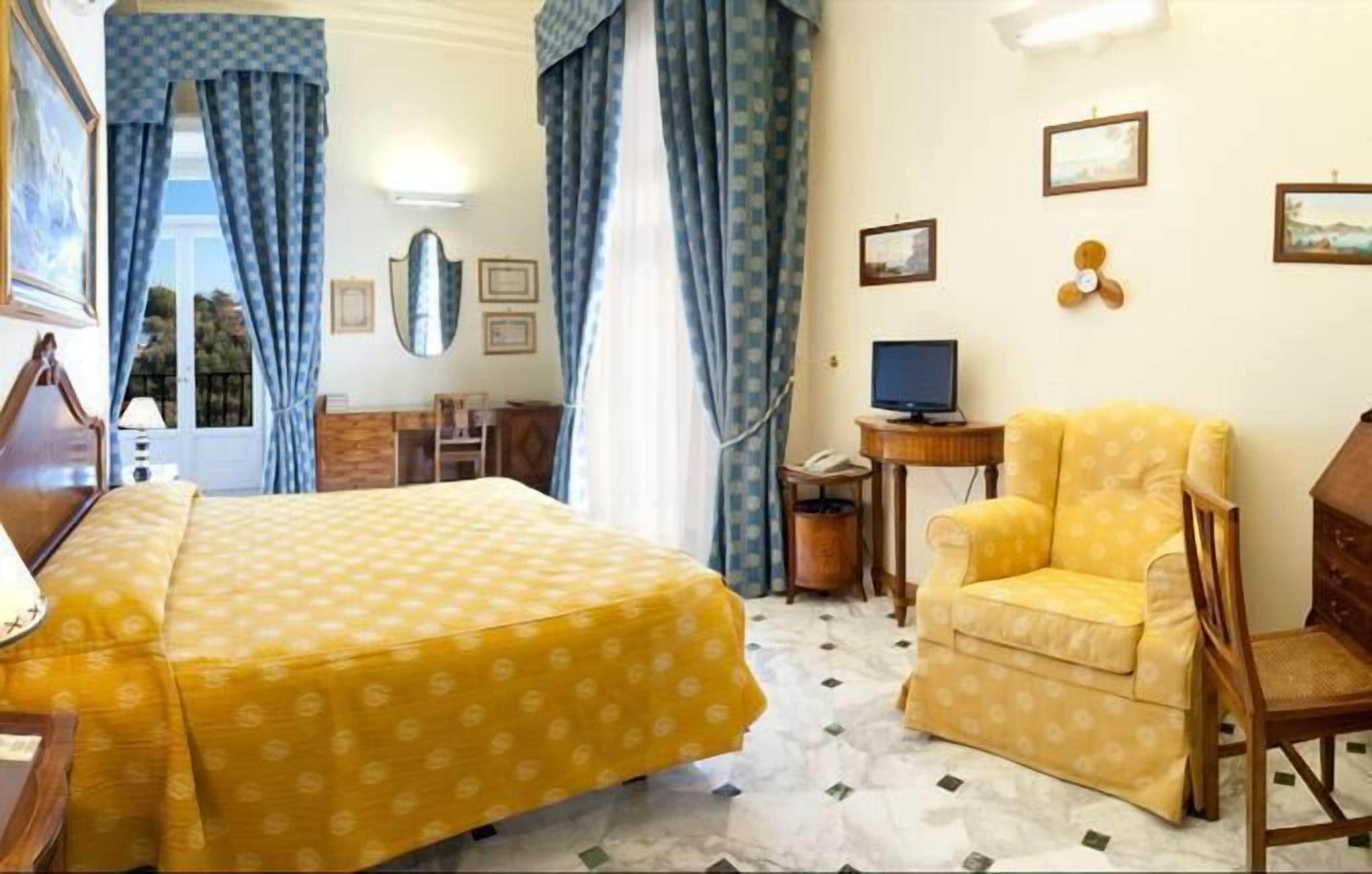 A hotel room with a bed, a chair, a desk with a computer, and a window with curtains.