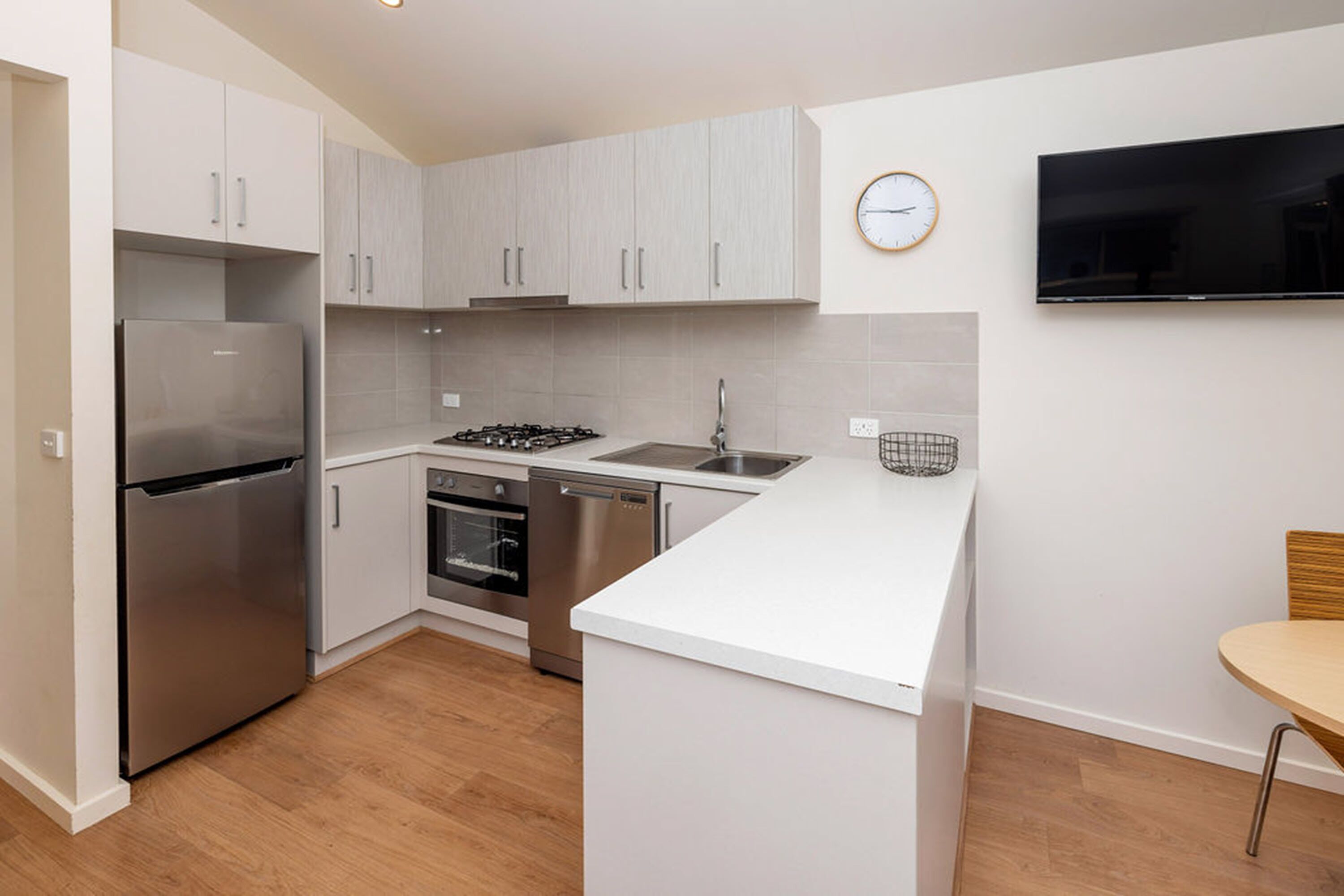 A modern kitchen with white cabinets, stainless steel appliances, a built-in oven, and a flat-screen TV mounted on the wall.