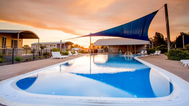 Una piscina con una vela de sombra azul, sillas de descanso y un edificio al fondo.