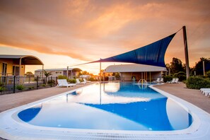 A swimming pool with a blue shade sail, lounge chairs, and a building in the background. - BIG4 Deniliquin Holiday Park (Deniliquin)