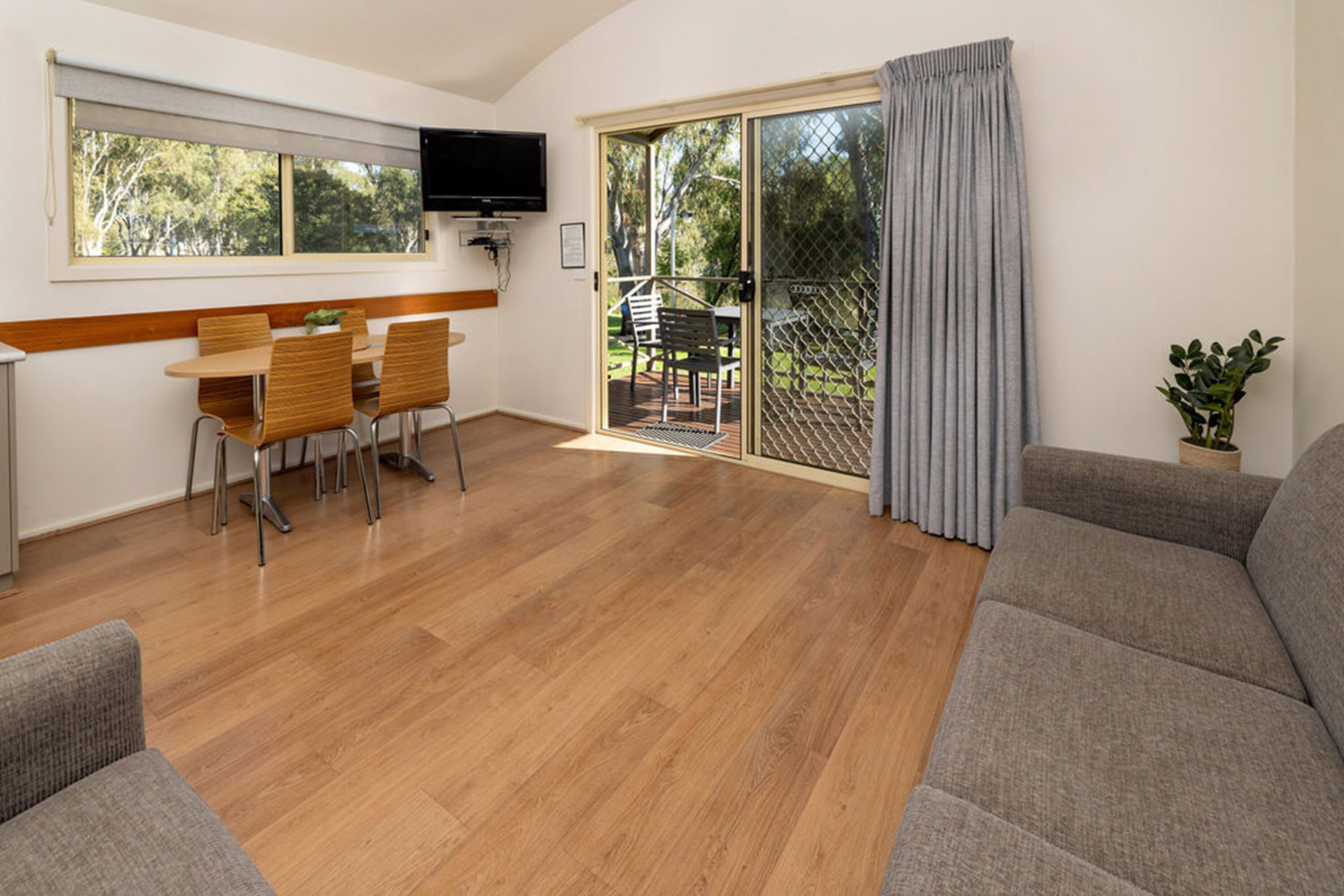A spacious living area with a wooden floor, a grey sofa, a dining table with chairs, a TV mounted on the wall, and a sliding glass door leading to an outdoor area.