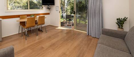 A spacious living area with a wooden floor, a grey sofa, a dining table with chairs, a TV mounted on the wall, and a sliding glass door leading to an outdoor area.