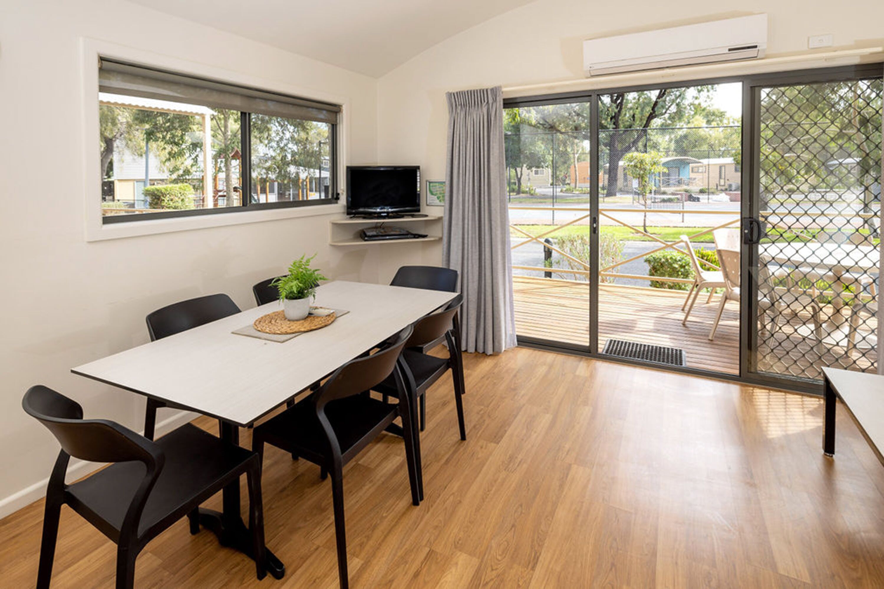A dining area with a white table, black chairs, a TV on a shelf, and a view of the outdoors through large sliding glass doors.