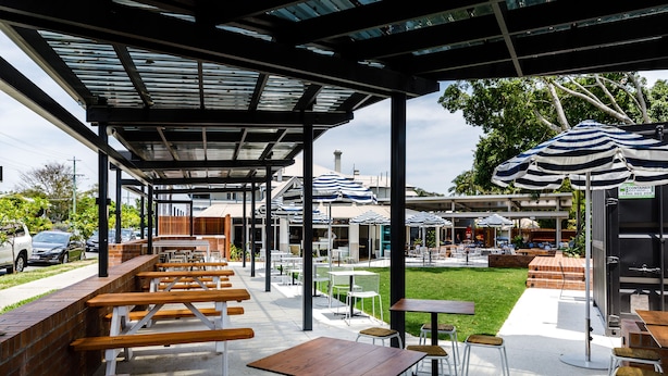 An outdoor patio area with wooden benches and tables, umbrellas, and a view of the street and greenery.