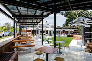 Un patio al aire libre con bancos y mesas de madera, sombrillas y vistas a la calle y áreas verdes.