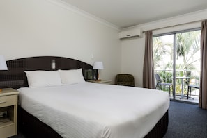 Habitación de hotel con una cama grande, una mesita de noche, una silla y un balcón con vistas a palmeras.