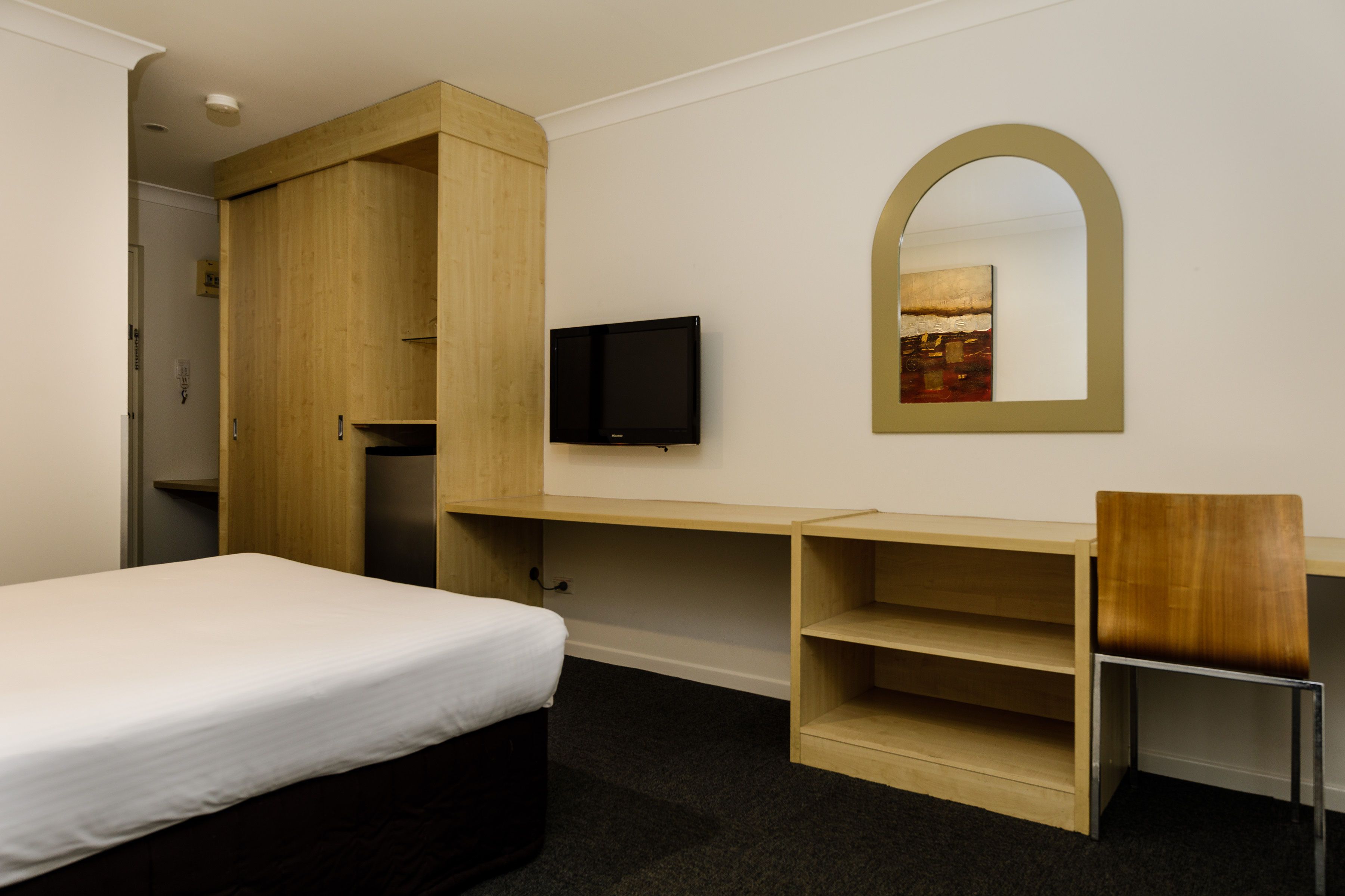 A hotel room with a bed, a wooden wardrobe, a TV, a desk with shelves, and a chair.