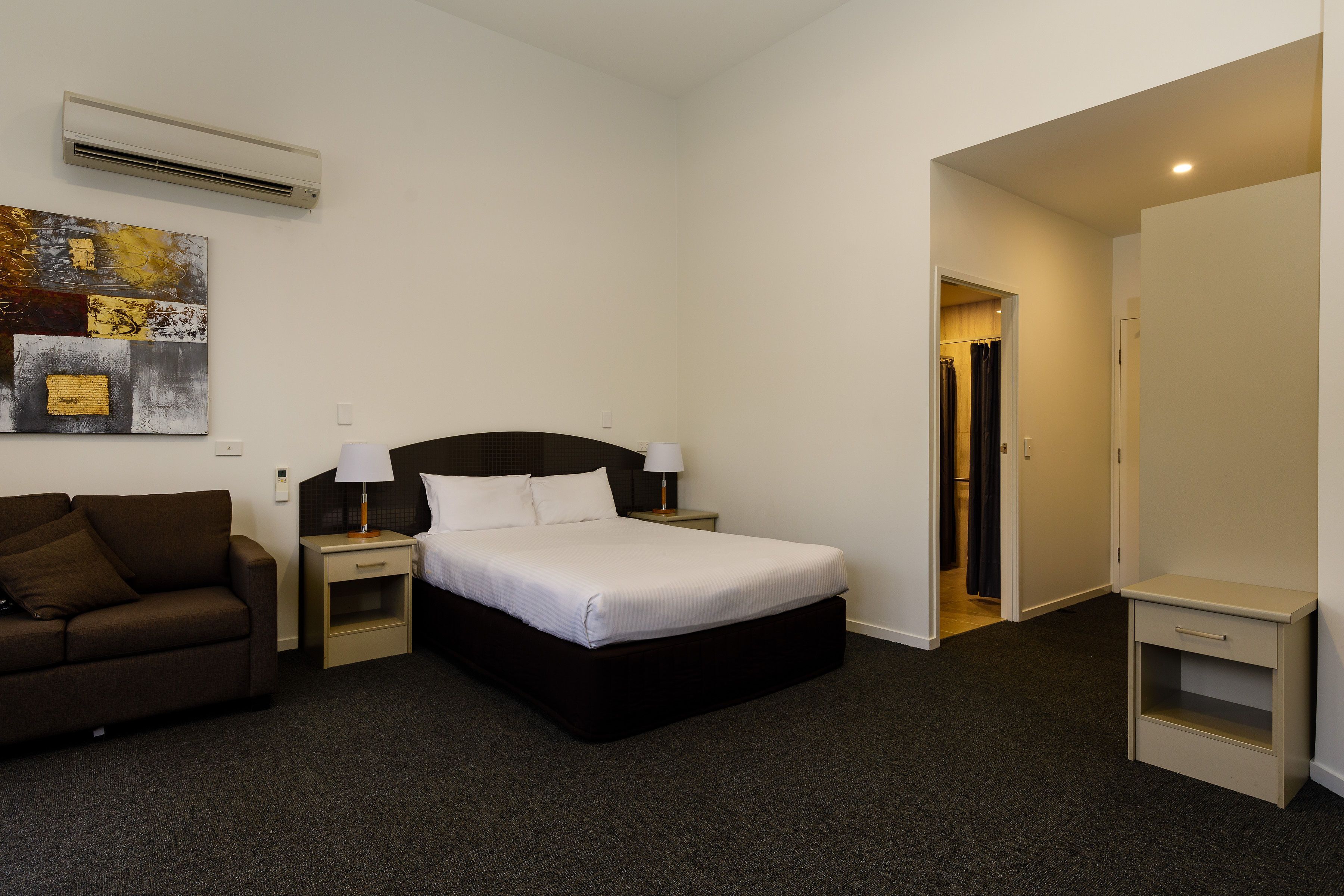 A hotel room with a bed, a nightstand, a lamp, a sofa, and a painting on the wall.