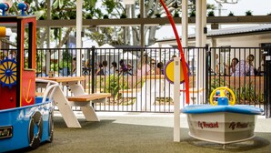 A children's play area with a small boat and a train, enclosed by a fence with benches and tables.