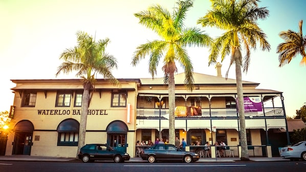Un edificio de dos pisos con el letrero "Waterloo Bar Hotel". Hay palmeras al frente y coches estacionados en la calle.