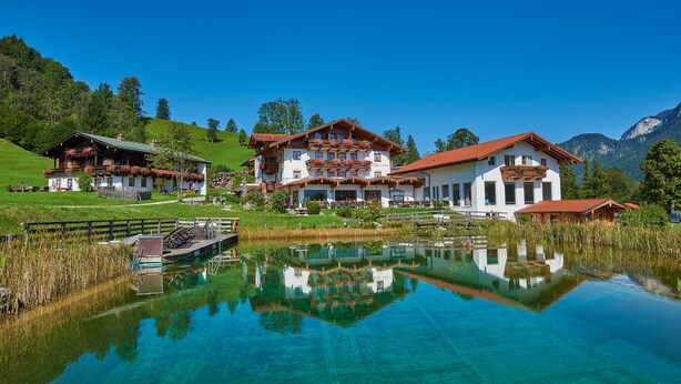 A large, multi-story building with balconies and a reflection in a clear body of water. - Naturhotel Reissenlehen (Bischofswiesen)